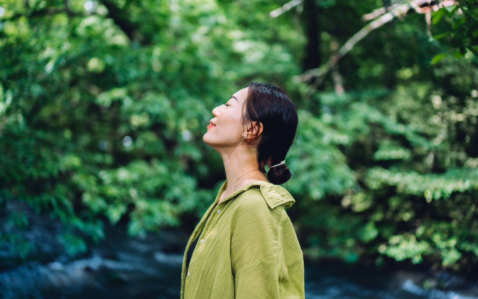 A woman in a green jacket is standing in the woods with her eyes closed.
