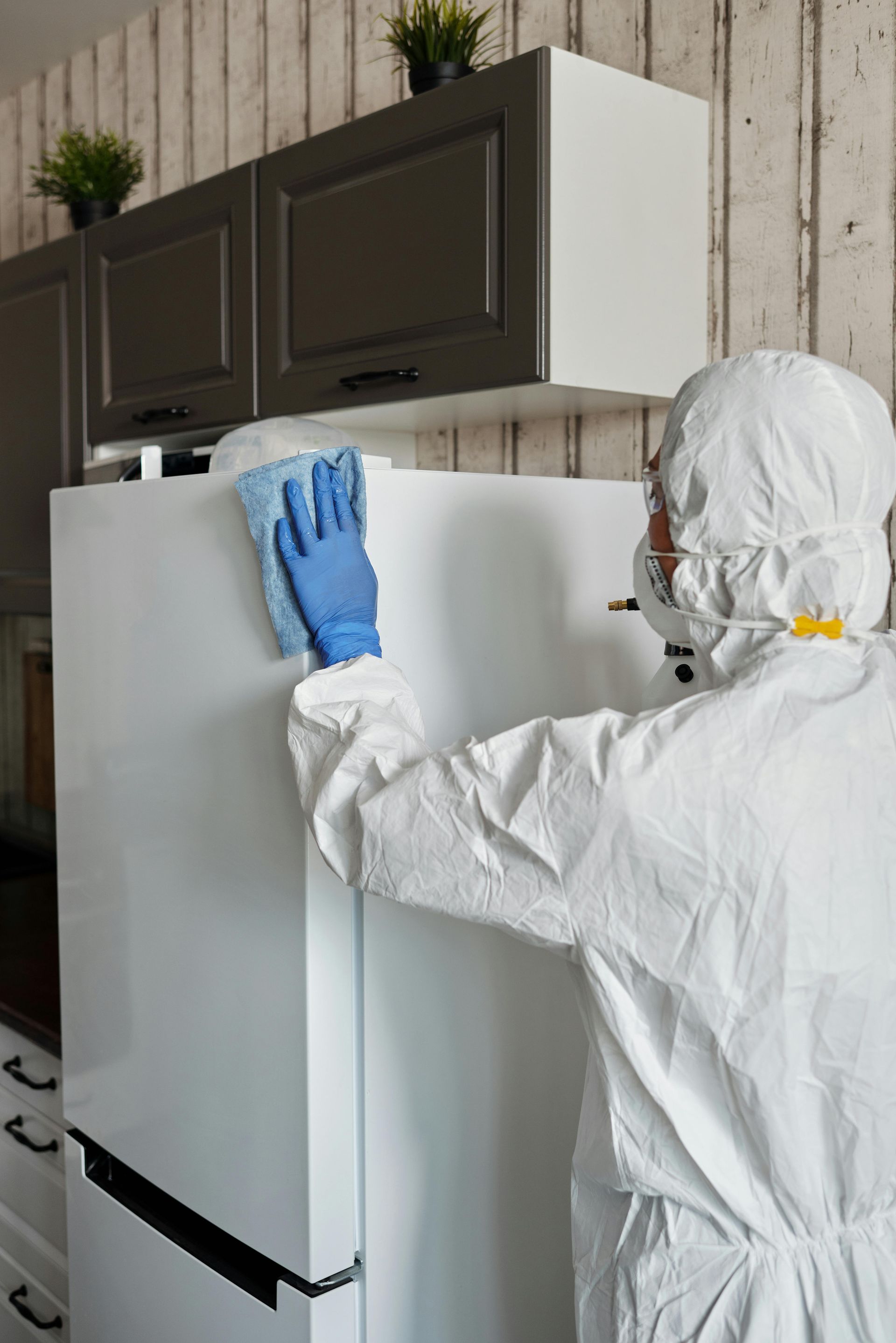 Person in hazmat suit cleaning a white refrigerator in a kitchen; wearing blue gloves and wiping with a blue cloth.