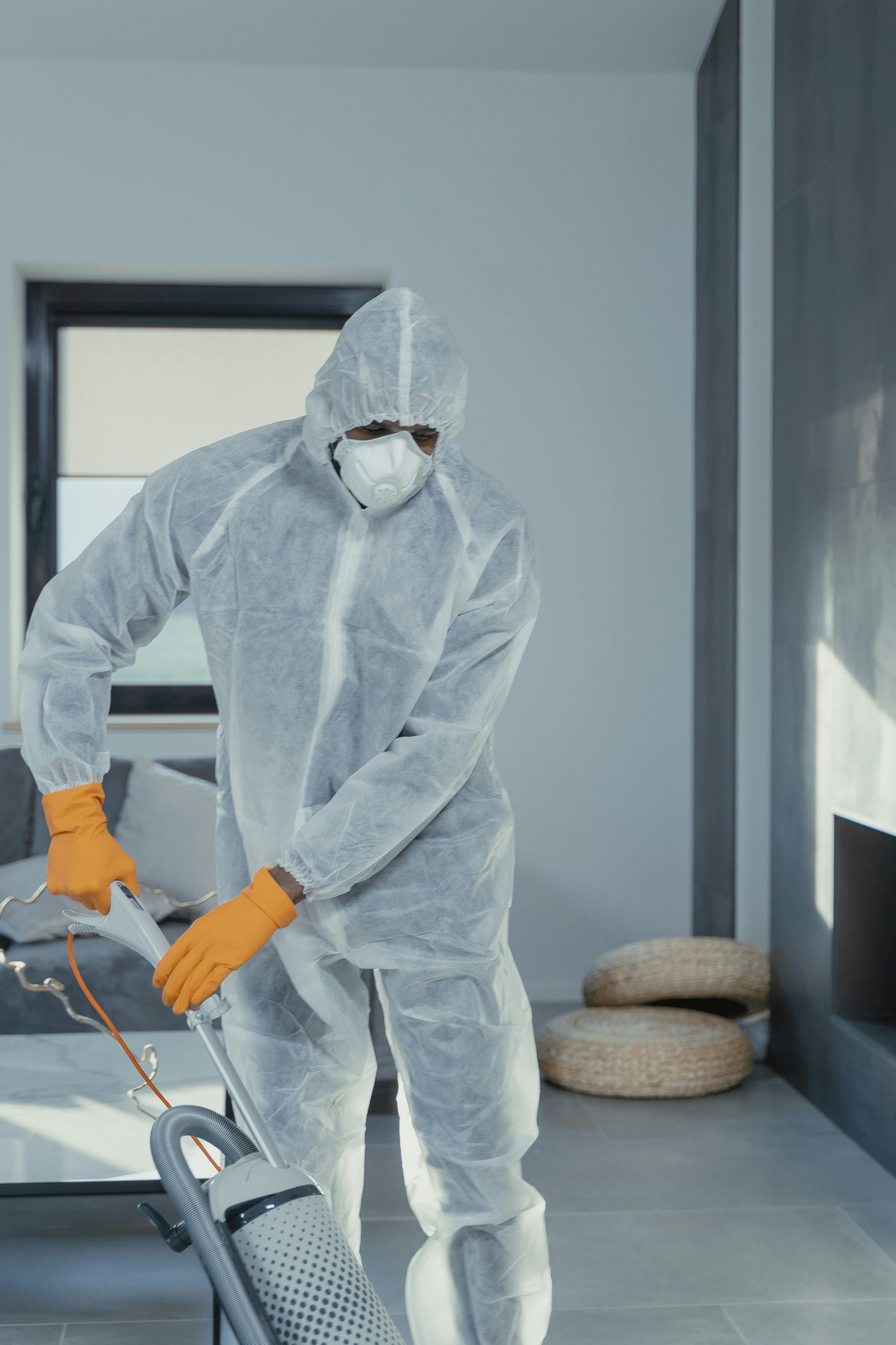Person in protective suit vacuums a tiled room, orange gloves, white suit and mask.