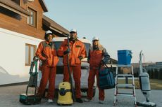 Three cleaning professionals posing with equipment outside a house; orange uniforms, smiling, sunny day.