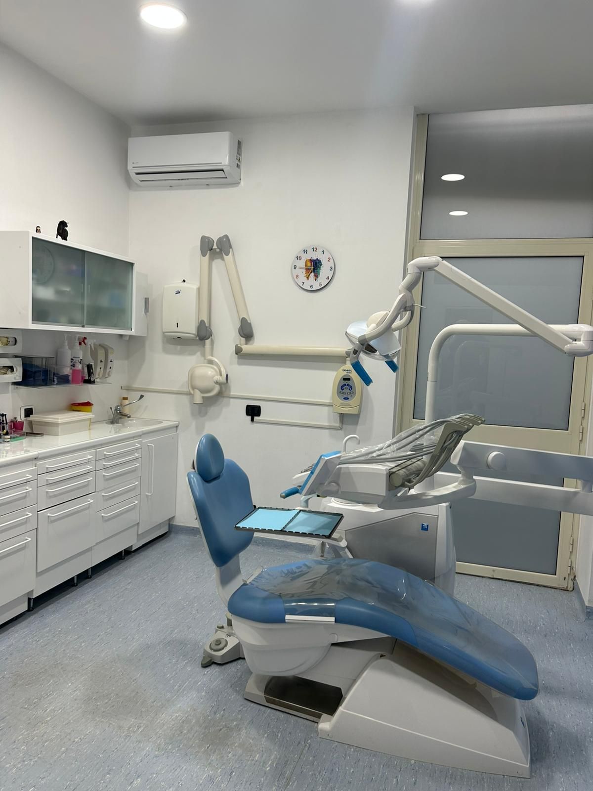 Sala operatoria odontoiatrica con poltrona blu, armadietti bianchi e attrezzatura odontoiatrica.