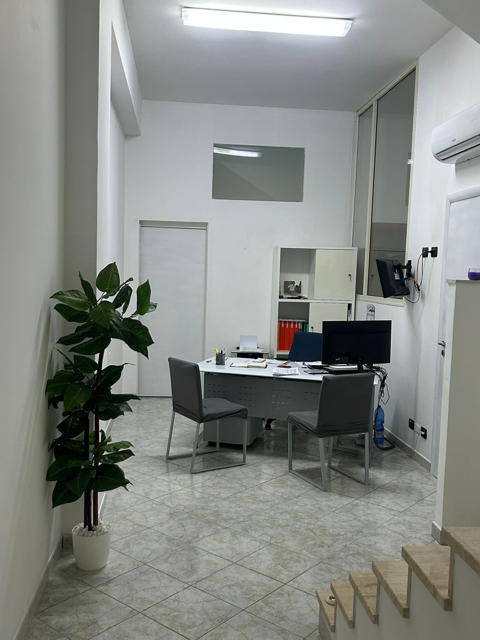 Interno di un ufficio con scrivania, sedie, una pianta e una scala. Pareti bianche e pavimento piastrellato.