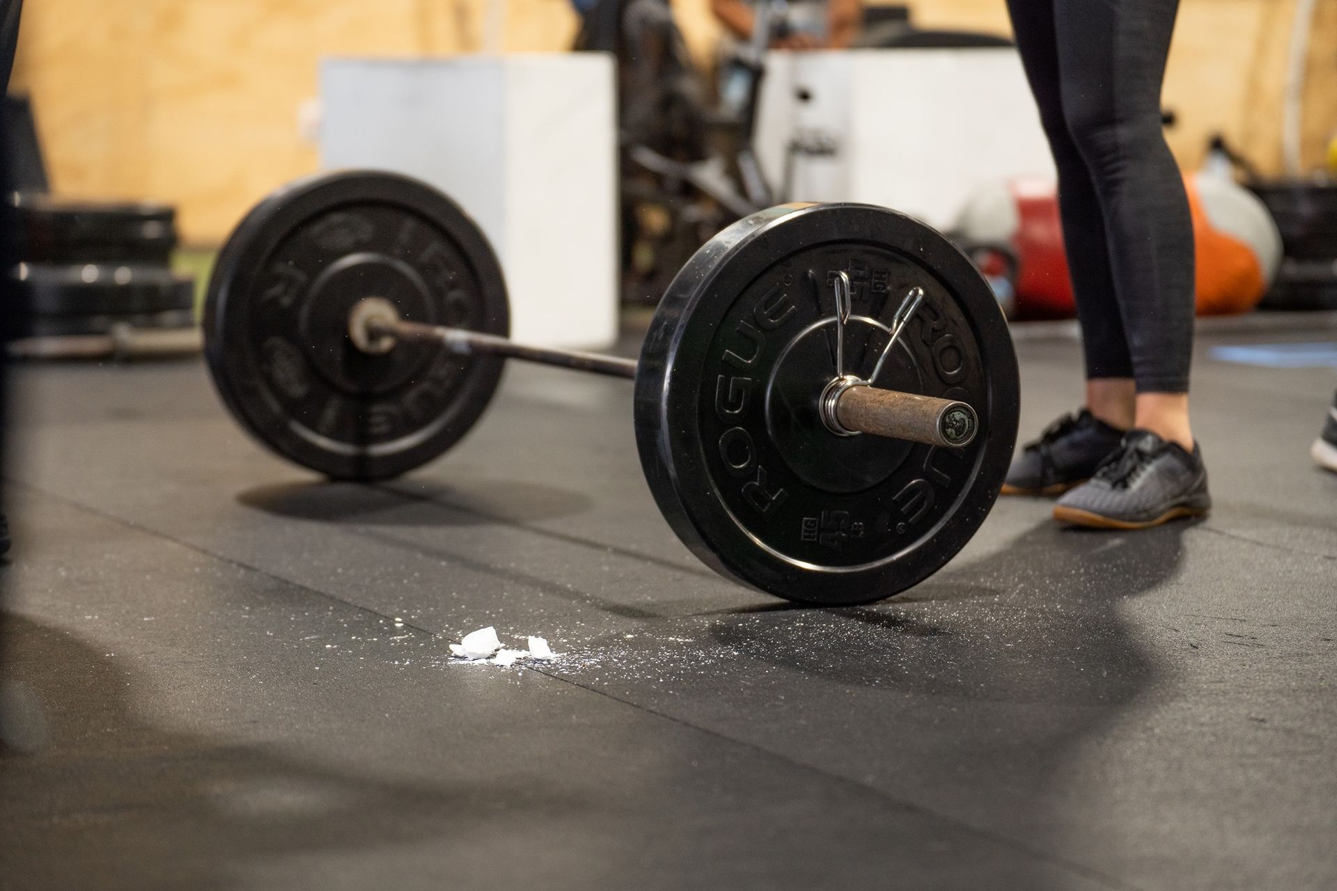 Barbell on a gym floor beside a person’s legs and shoes, with chalk dust nearby at the best crossfit gym in Fort Walton Beach, FL