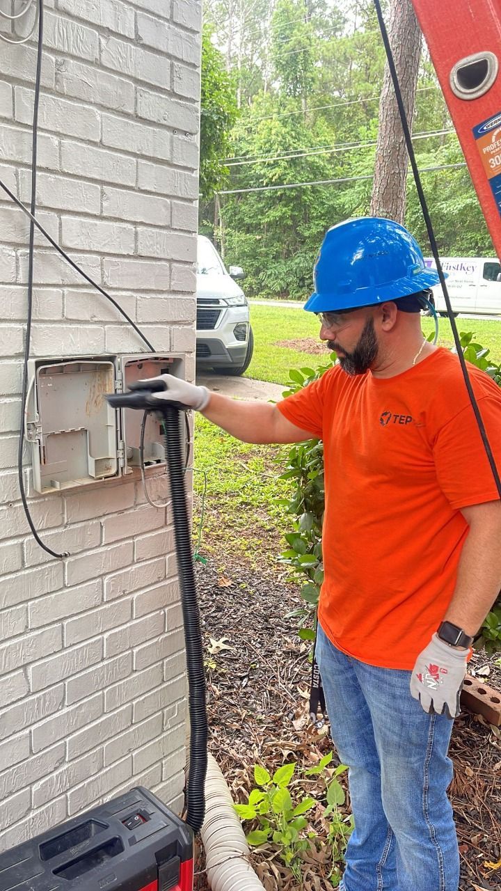Man in orange shirt and blue hardhat works on a wall-mounted box on a brick building.