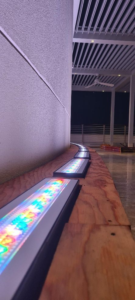 A curved row of colorful LED lights on a wooden surface next to a white wall and a pergola.