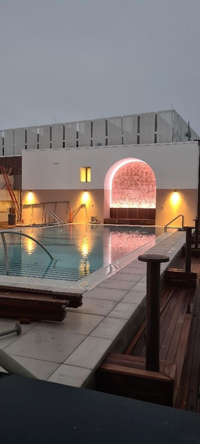 A rooftop pool at dusk, illuminated by warm lights. White architecture with archway and reflecting pool.