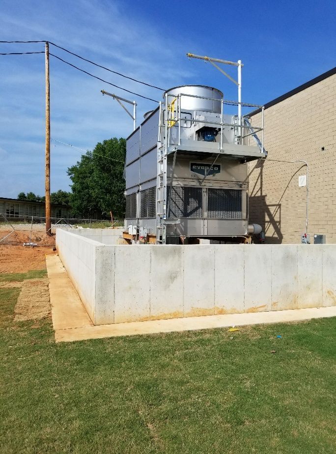 A metal cooling tower on a concrete base next to a brick building under a blue sky.