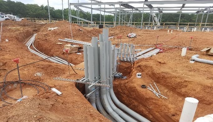 Construction site with grey electrical conduits in trenches; building frame in background.