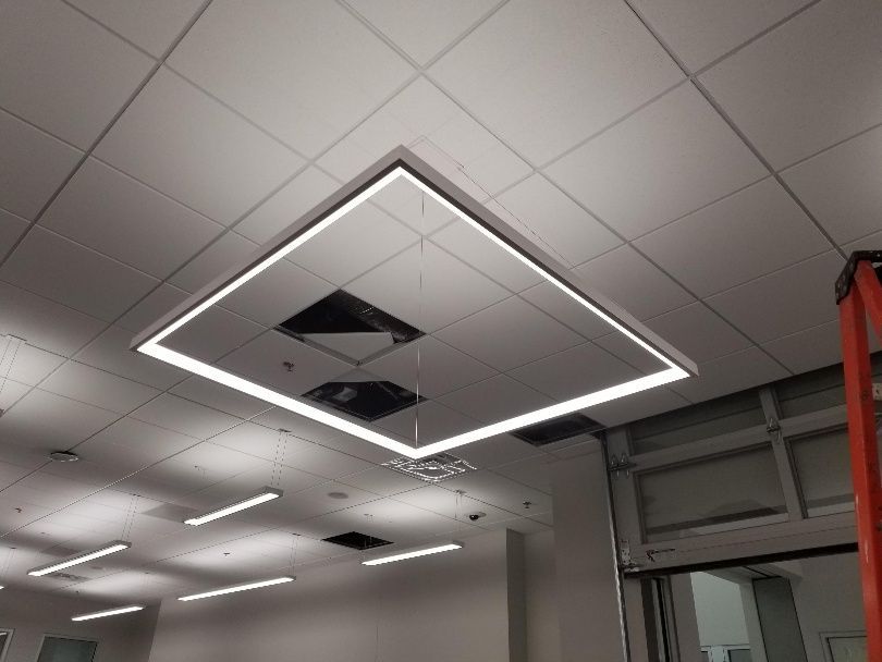 Square LED light fixture suspended from a white ceiling, with fluorescent lights in the distance and a ladder on the right.