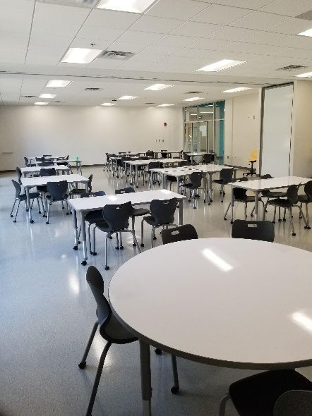 Empty school cafeteria with tables and chairs, white walls, bright lighting.