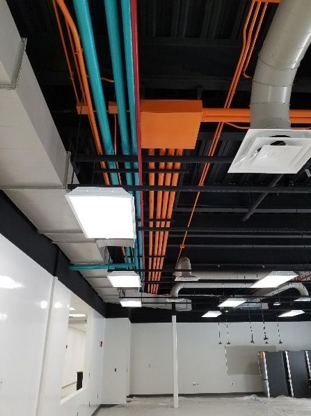 Interior view of a ceiling with exposed colorful pipes and ducts in a room with white walls and fluorescent lights.