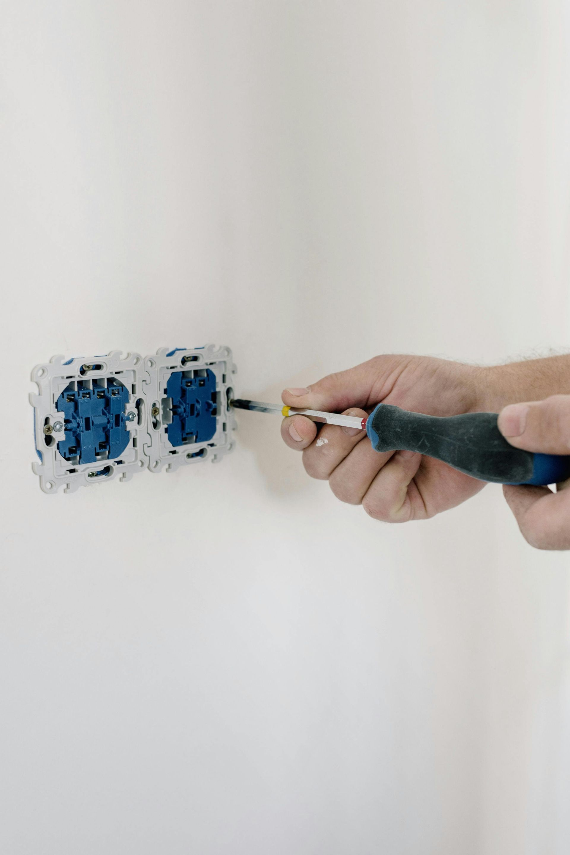 Hand with screwdriver connecting a wire to a blue electrical outlet set against a white wall.