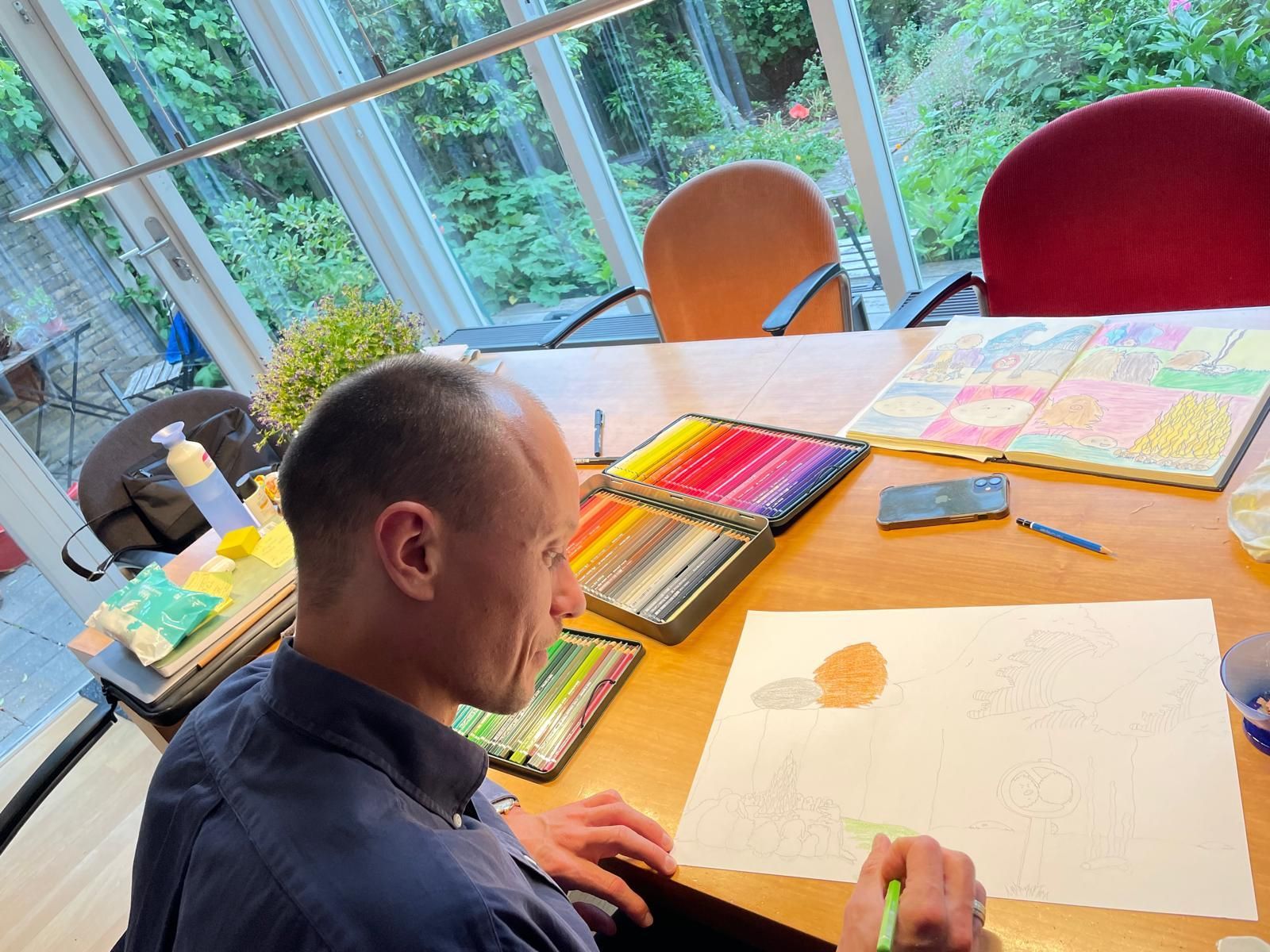 Een man tekent met kleurpotloden aan een tafel. Op de tafel liggen ook diverse kunstbenodigdheden en kleurrijke kunstwerken.