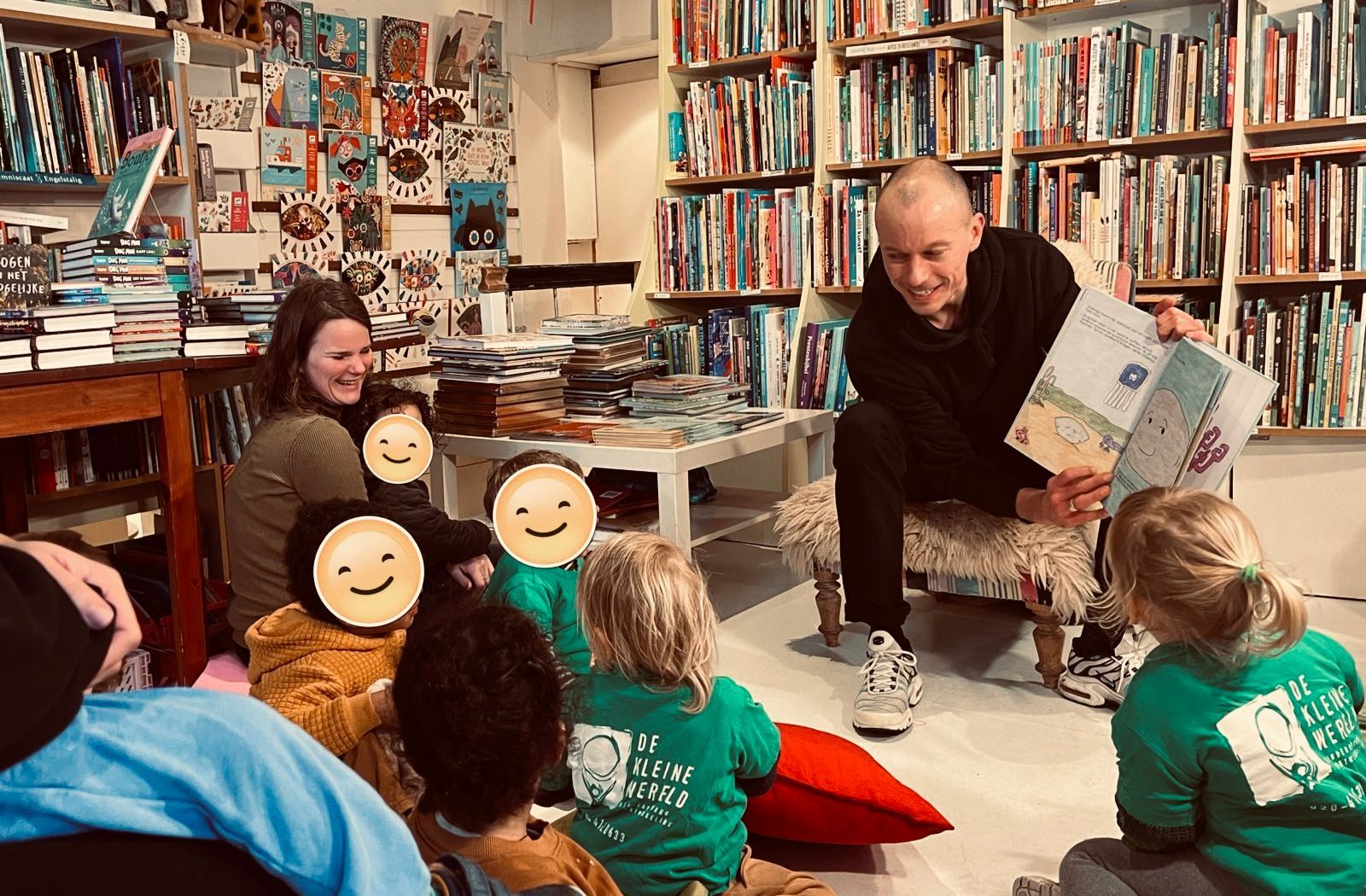 Een man leest een boek voor aan kinderen in een boekwinkel. Op de achtergrond zijn boekenplanken te zien, samen met een vrouw en een rood kussen.