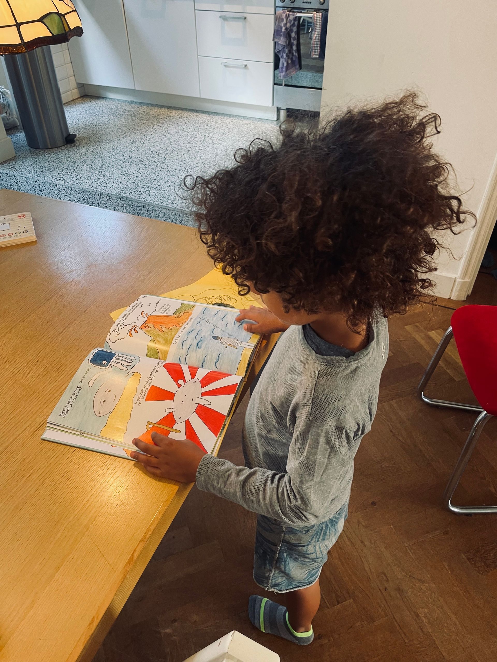 Kind met krullend haar leest een boek aan een tafel met een mozaïekvloer.