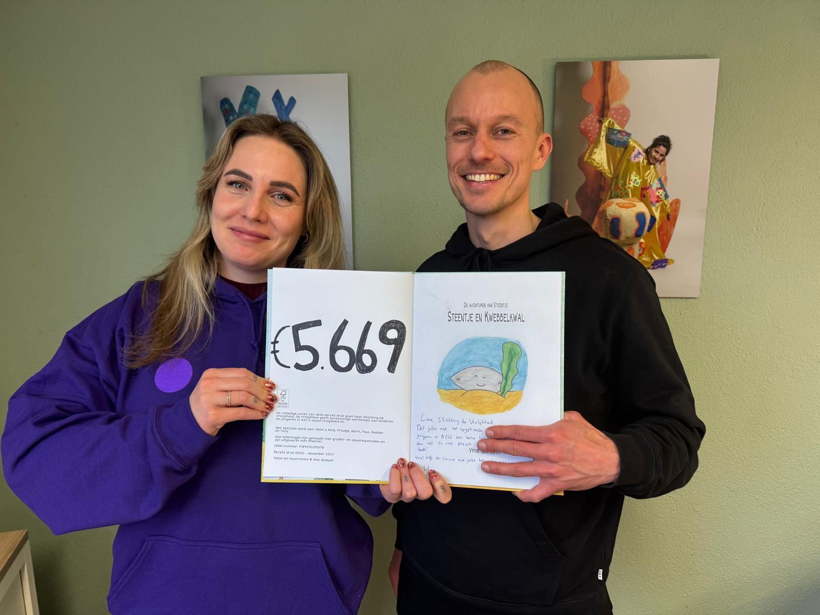 Twee mensen houden een boek vast met het opgehaalde bedrag van 5669 euro. Ze staan ​​lachend voor een muur met schilderijen.