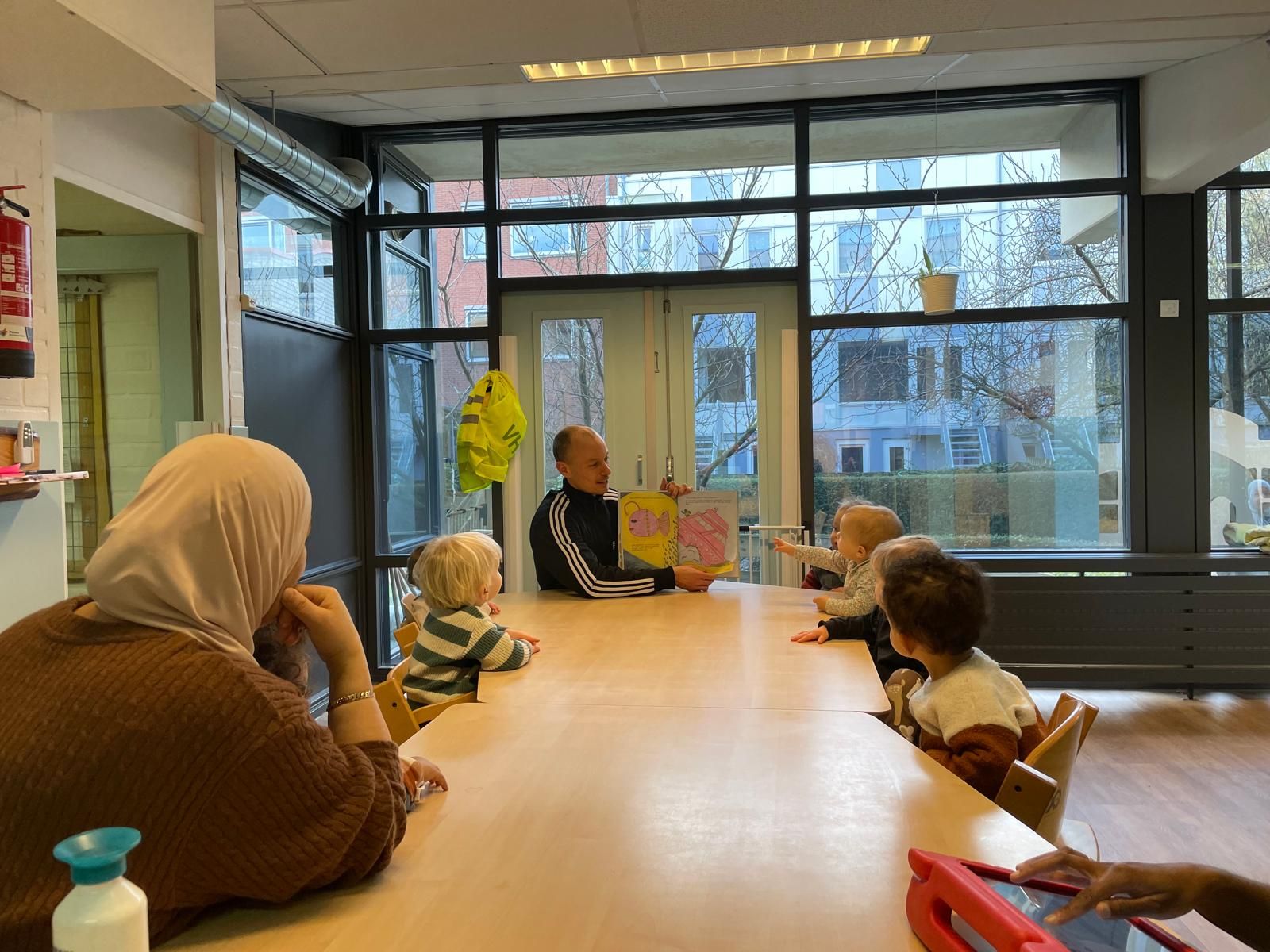 Een leraar leest voor aan kinderen aan een tafel; een andere volwassene kijkt toe. Vanuit de ramen heb je uitzicht naar buiten.