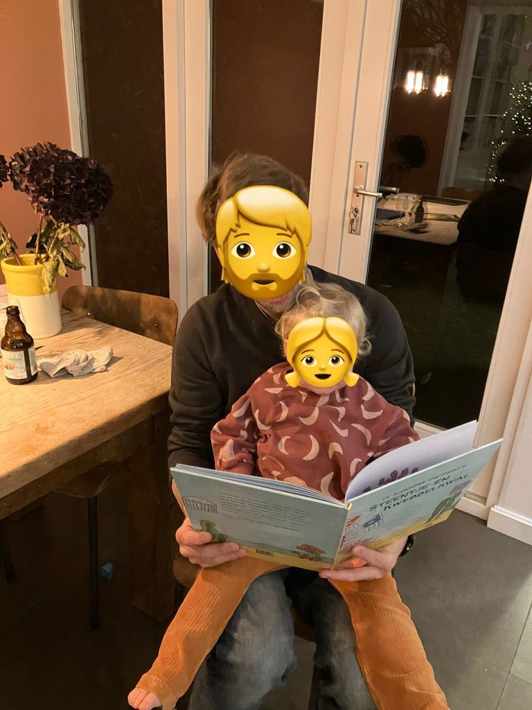 Persoon met kind zittend, lezend. Binnenshuis, zachte verlichting, persoon in zwarte trui, kind in gebloemd overhemd.