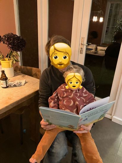 Een persoon houdt een kind vast, beiden kijken naar een open boek. Binnen, warm licht.