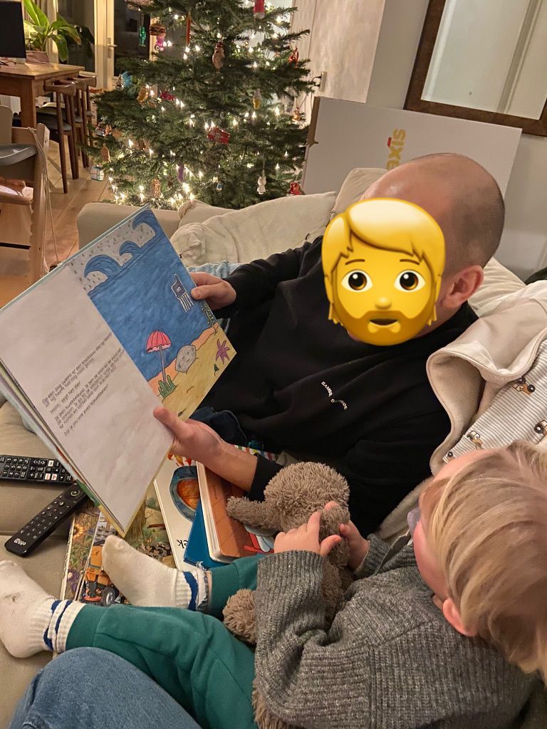 Een persoon leest een boek voor aan een kind op de bank, met een kerstboom op de achtergrond.