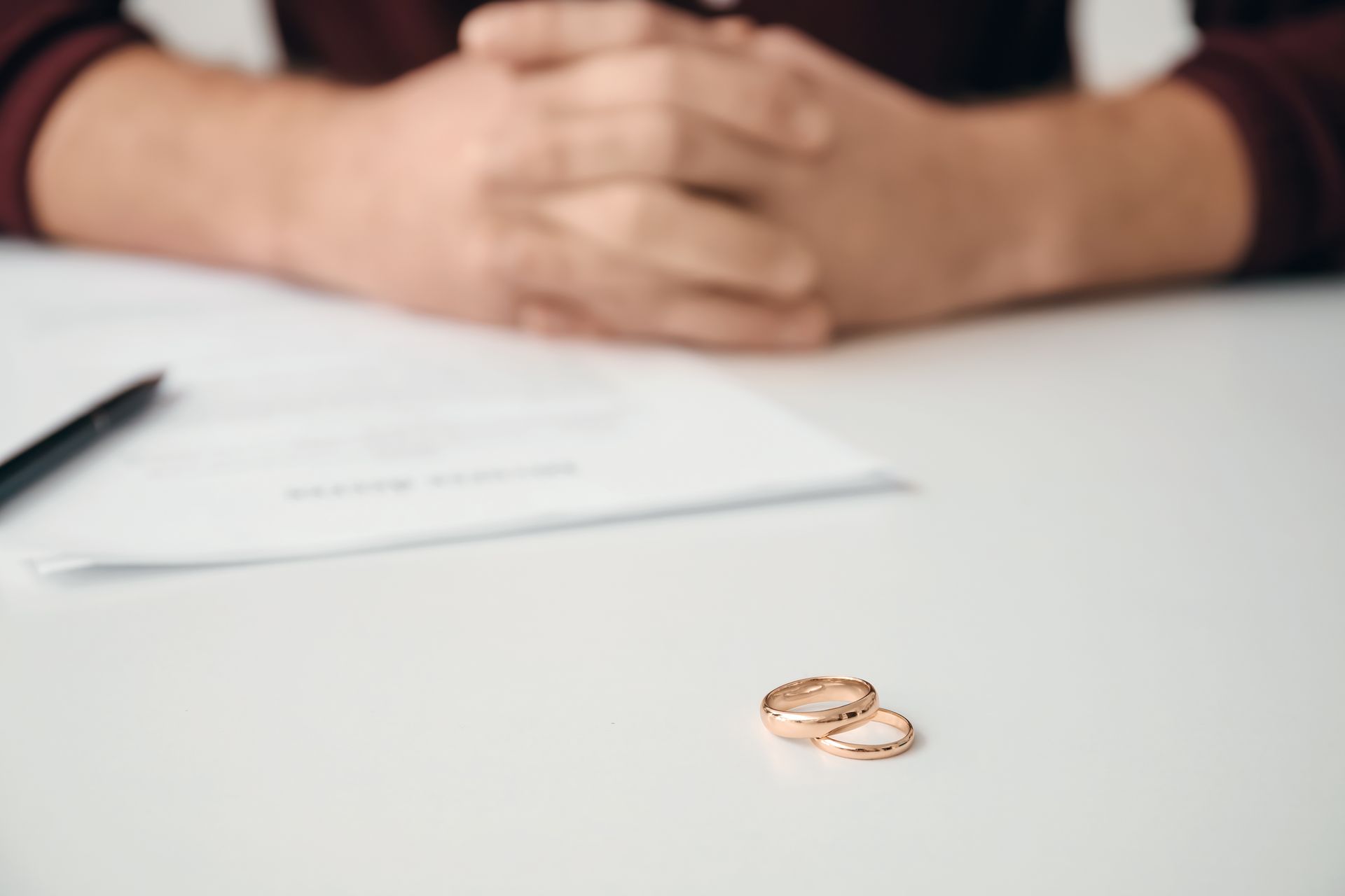 A woman is sitting at a table with her hands folded and a pair of wedding rings on the table.