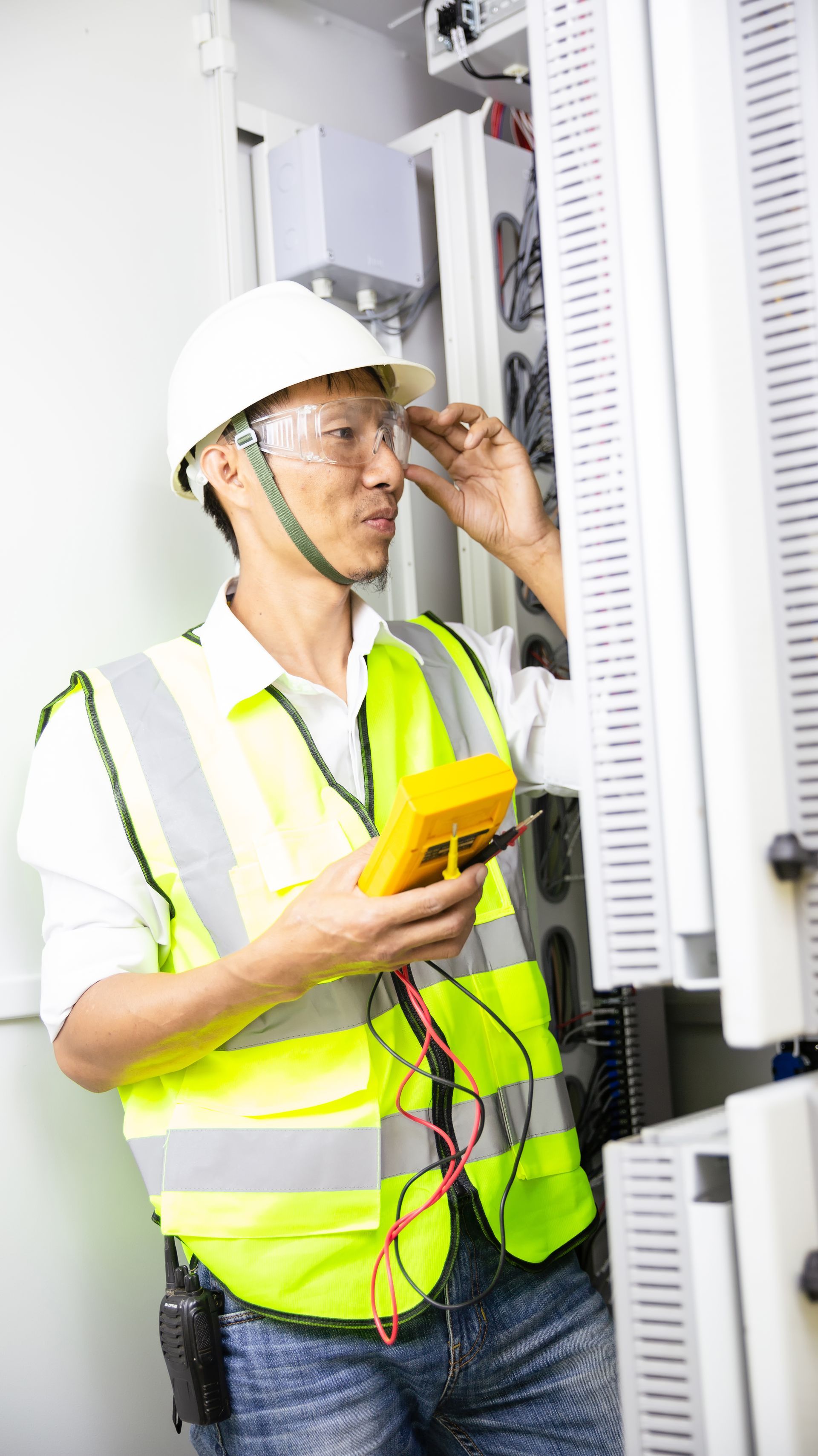 A man in a hard hat and safety vest is using a multimeter in a server room.