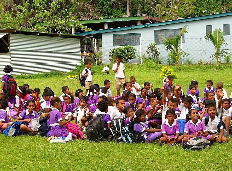 Schoolchildren in purple uniforms seated on grass, school buildings in background.
