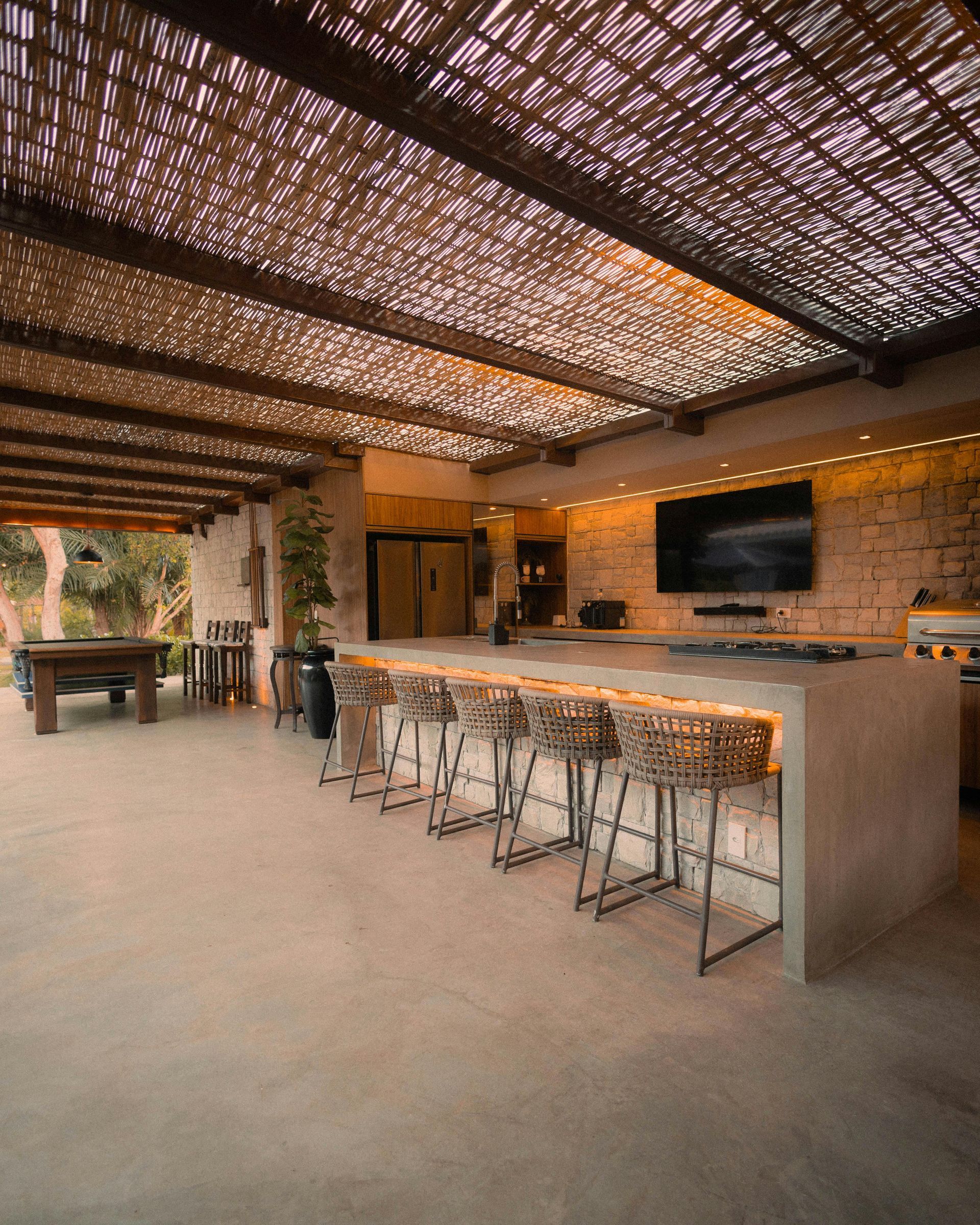 thatch roof outdoor cement bar with wicker-backed chairs facing grill area and large outdoor tv on house stone wall