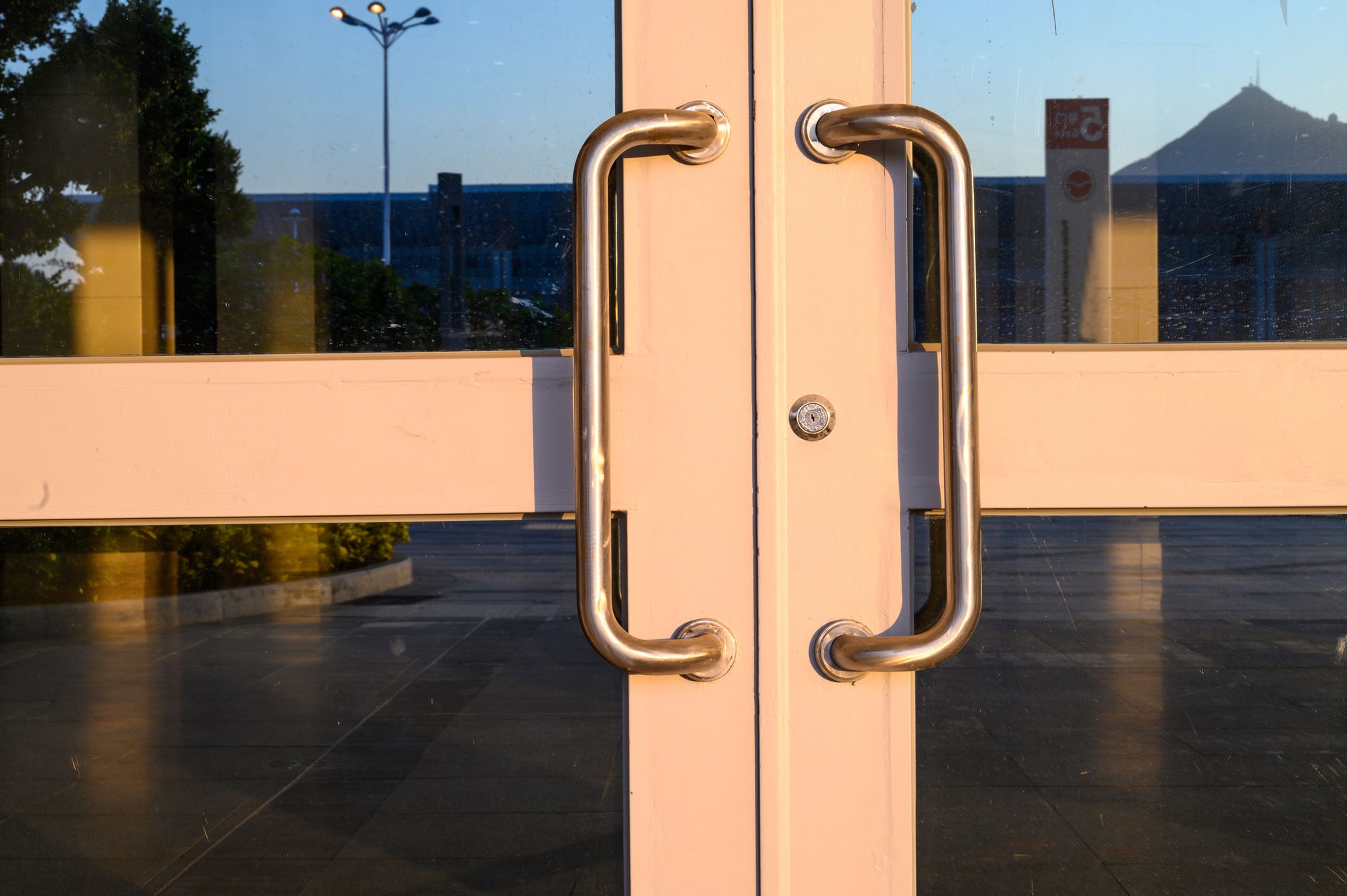 Glass commercial door with metal handles and keyhole, reflecting trees and clear sky.