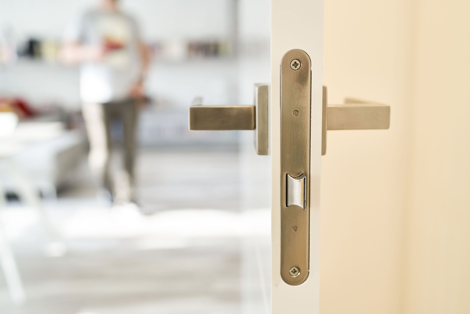 Close-up of a brushed metal door handle & lock on a white door, with a blurred person in the background.