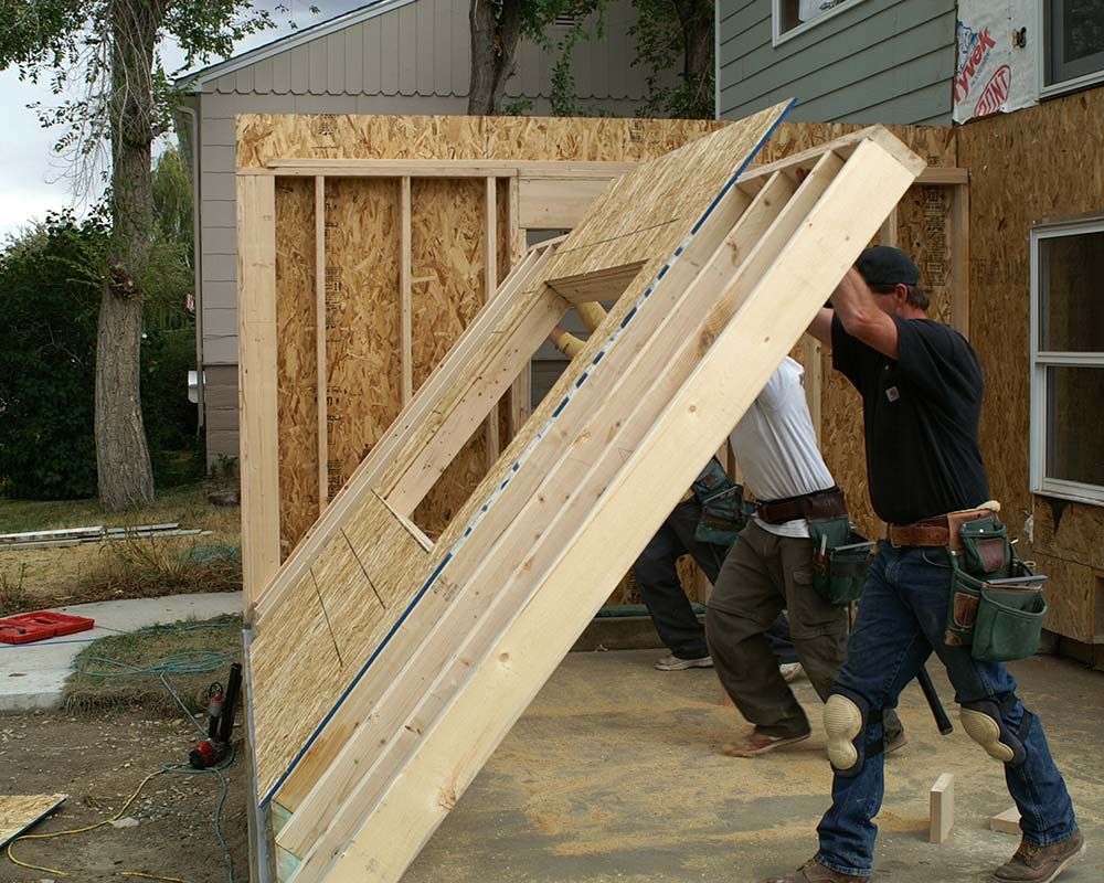 Construction workers lift a wall frame, likely for a house addition, outdoors on a sunny day.