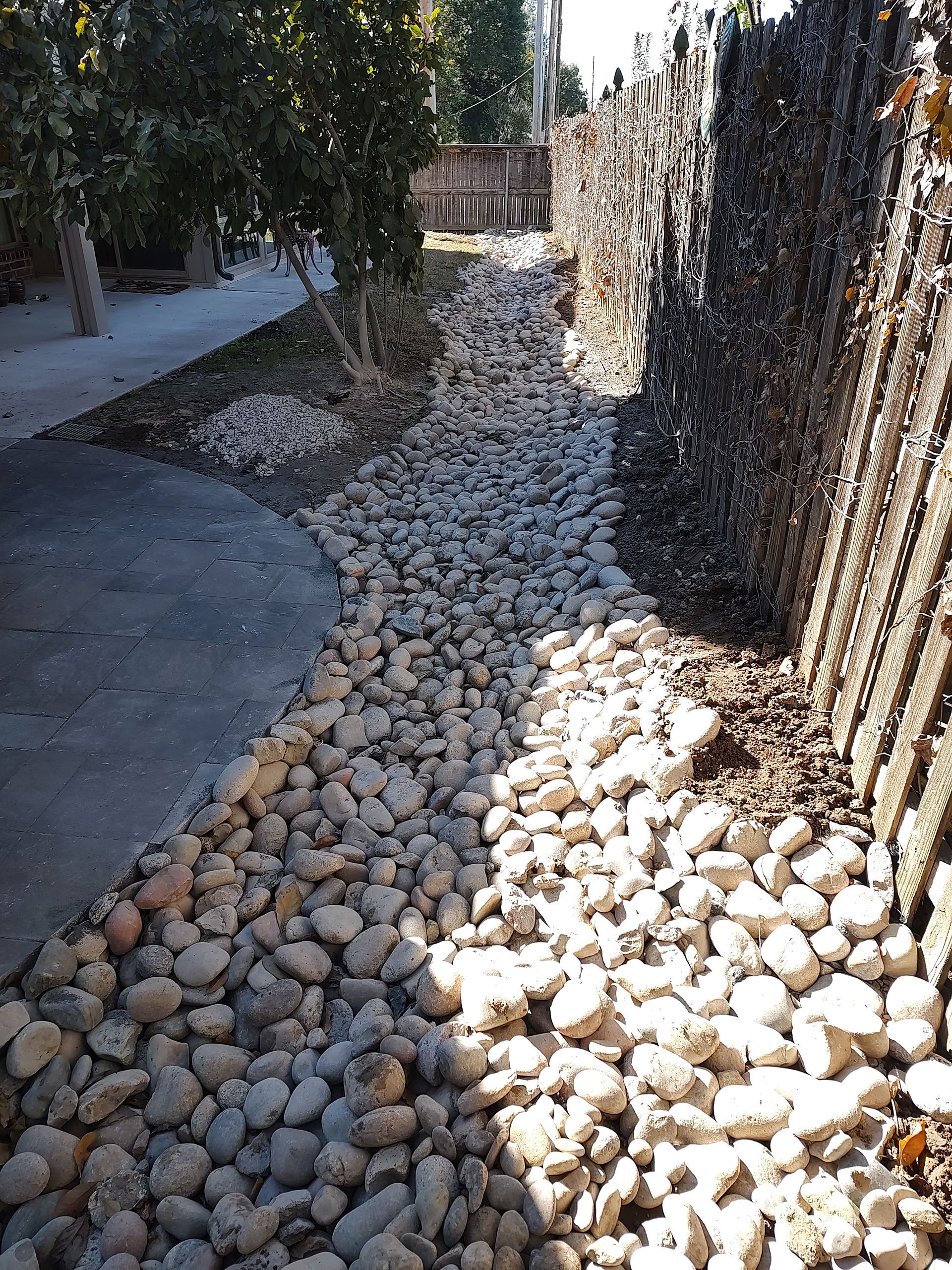 A dry riverbed of rocks next to a concrete path and a stone wall.