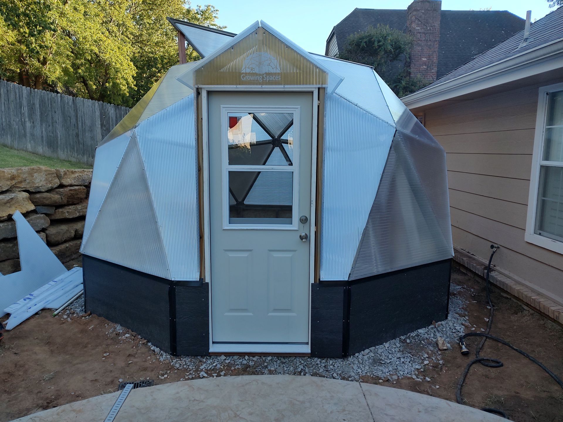 Geodesic dome structure with a door, made of corrugated metal and black panels in a backyard.