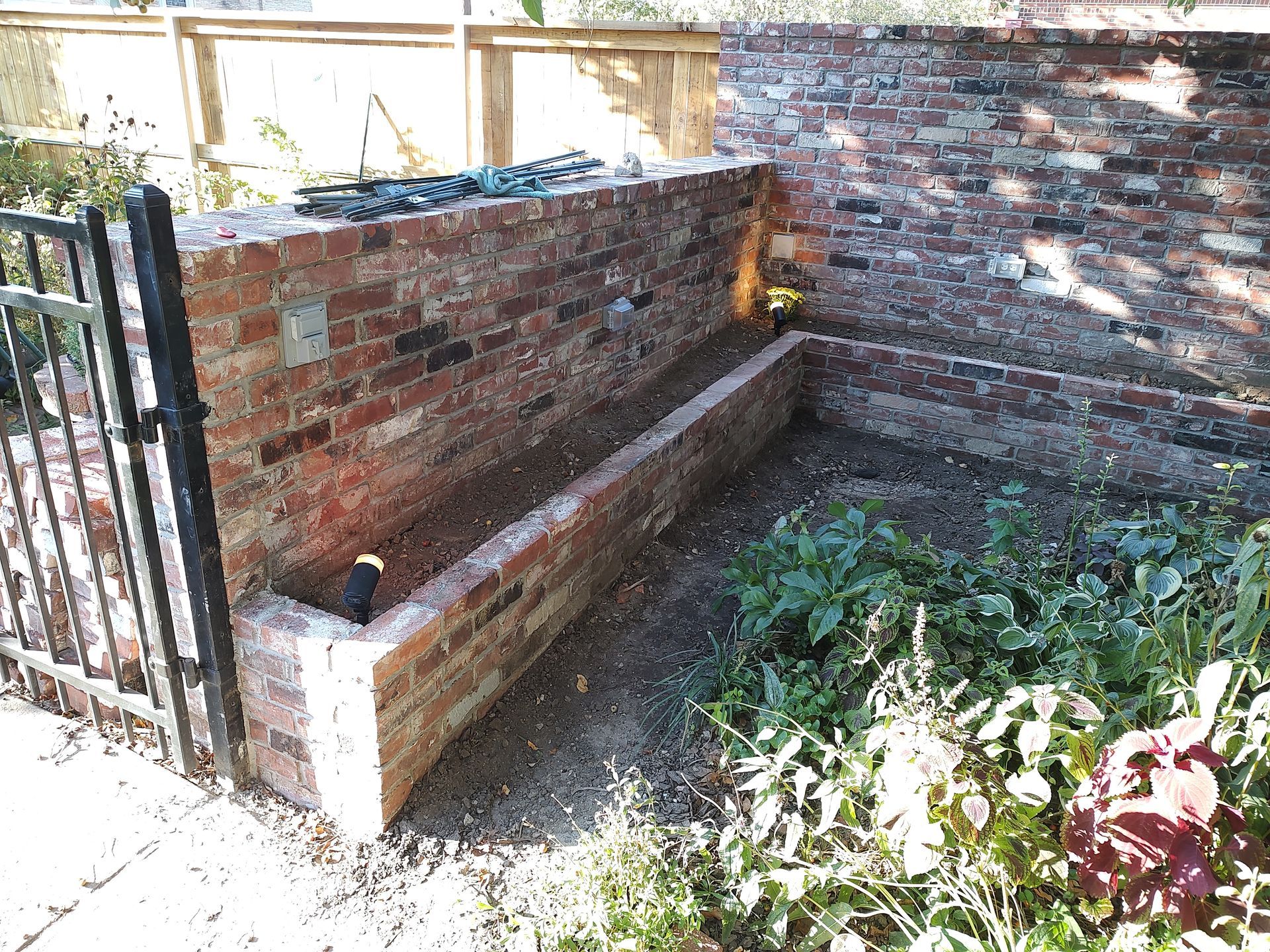 Brick planter box next to a brick wall and black metal gate. Green plants grow inside.