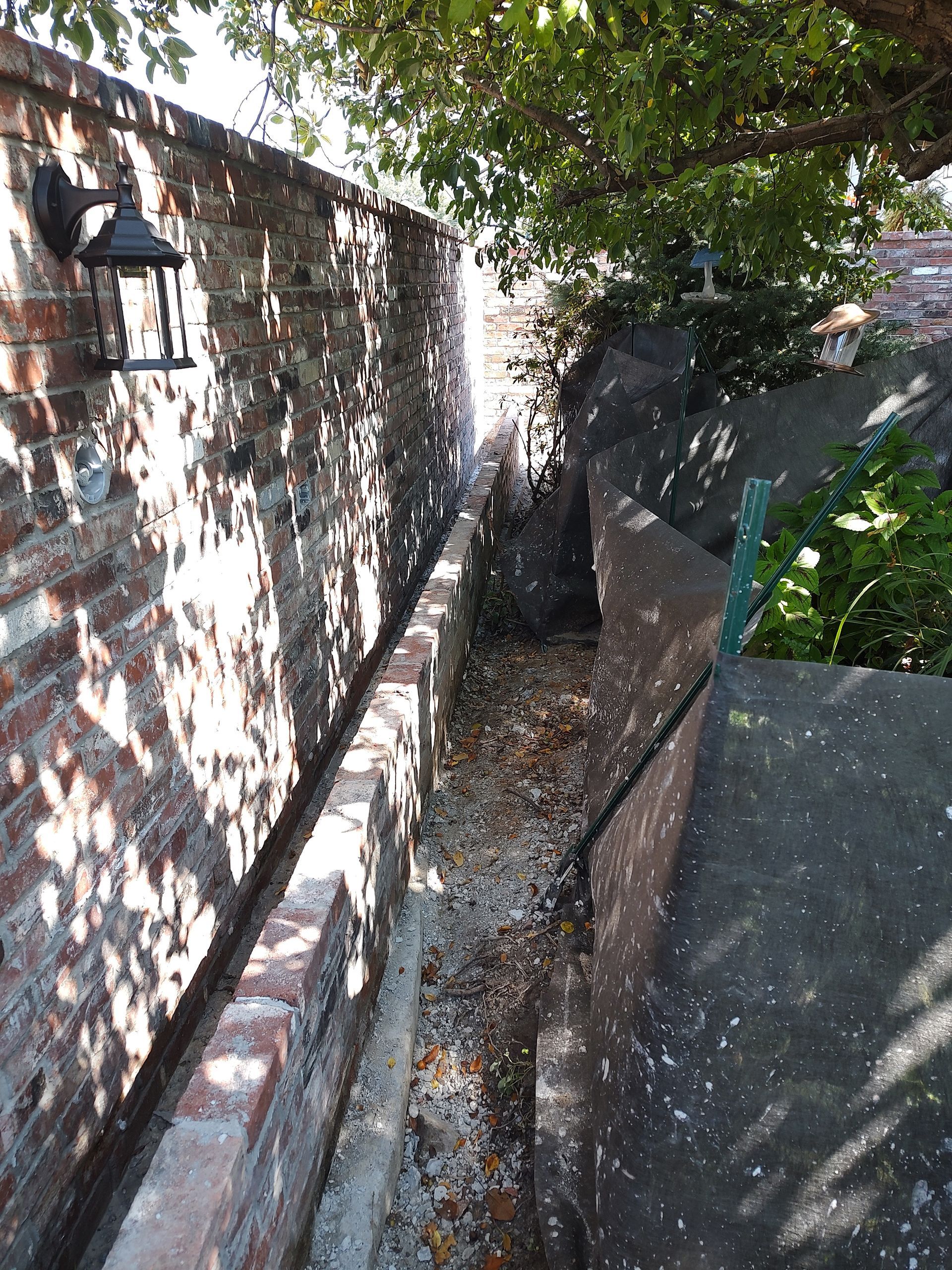 Narrow alley between a brick wall with a lantern and a concrete structure, filled with leaves.