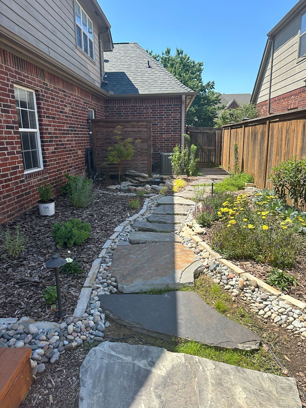 Stone path through a landscaped yard with plants and a wooden fence, next to a brick building.