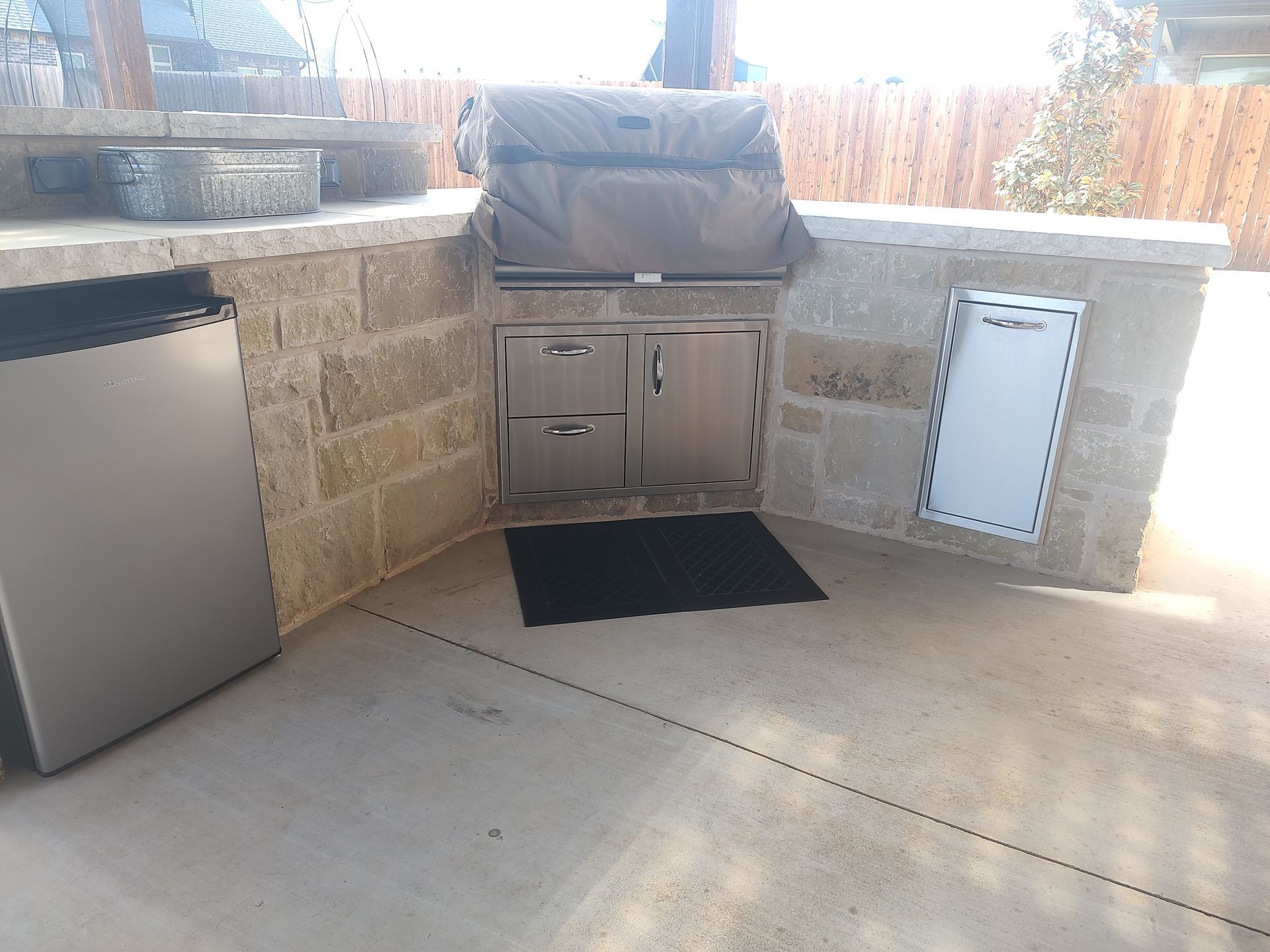 Outdoor kitchen with stainless steel grill, cabinets, and built-in trash receptacle. Stone counter and gray concrete patio.