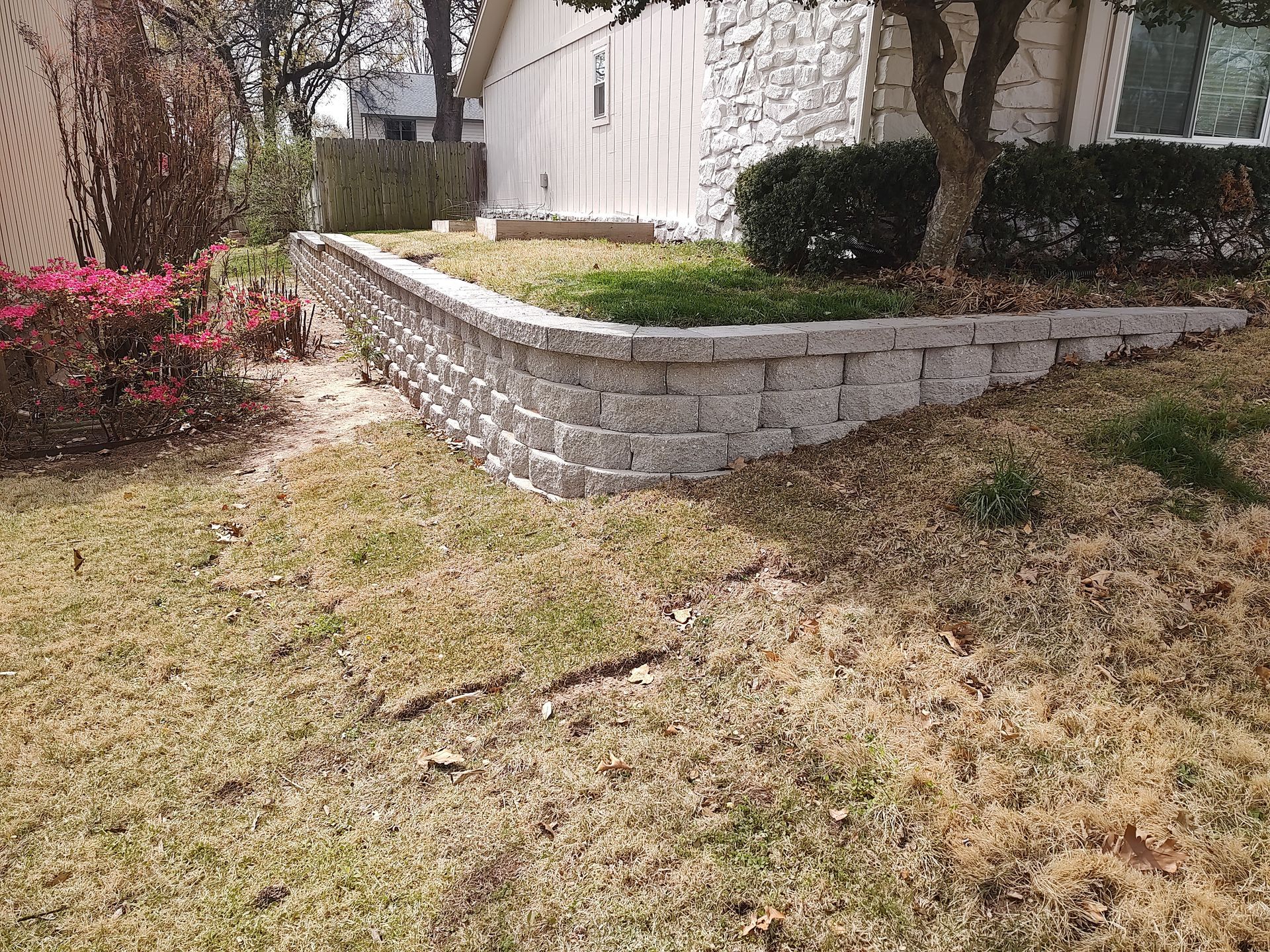 A retaining wall made of gray blocks borders a lawn; a house and small tree are in the background.