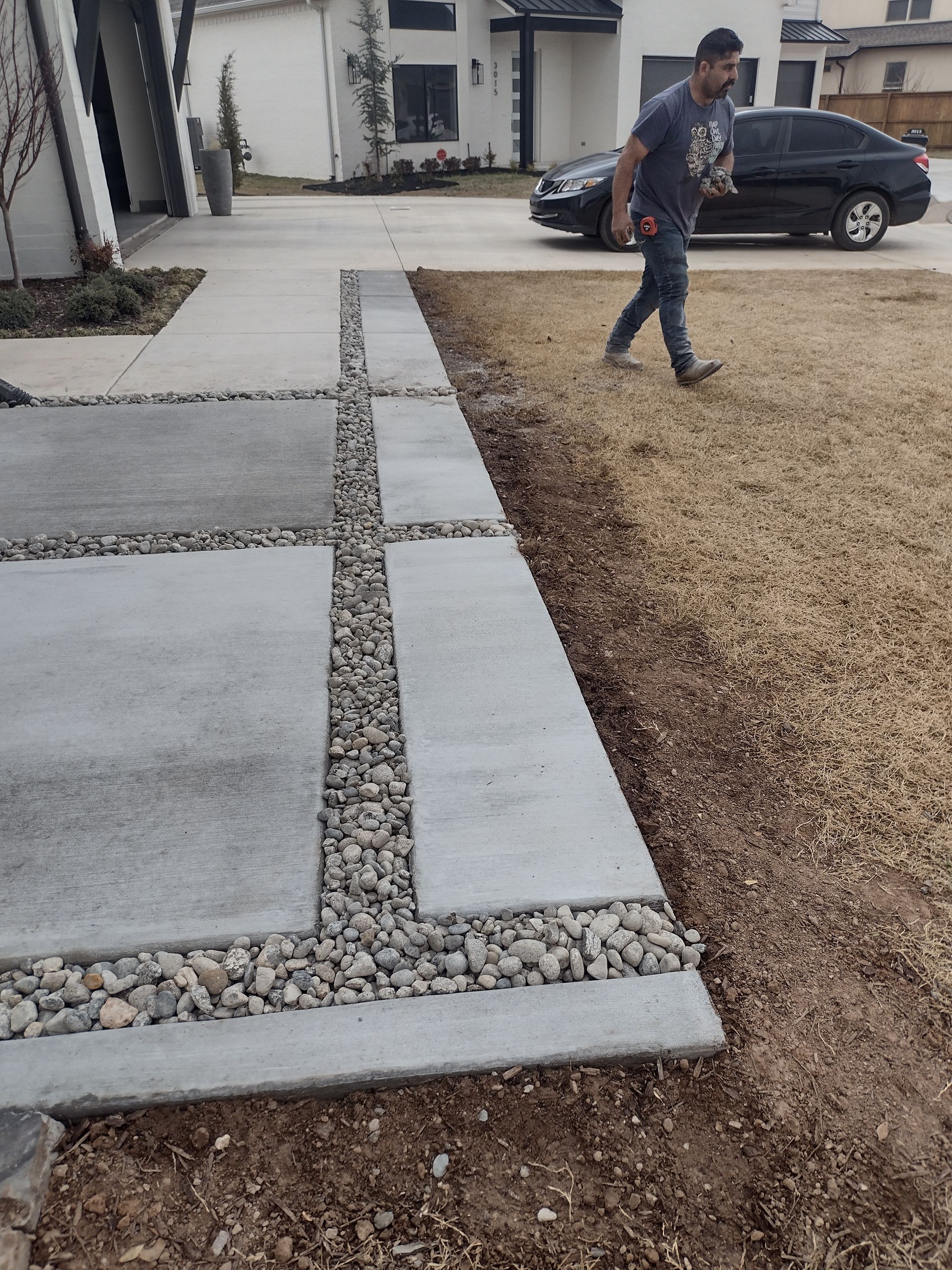 Man walking near concrete walkway with gravel accents; brown lawn.