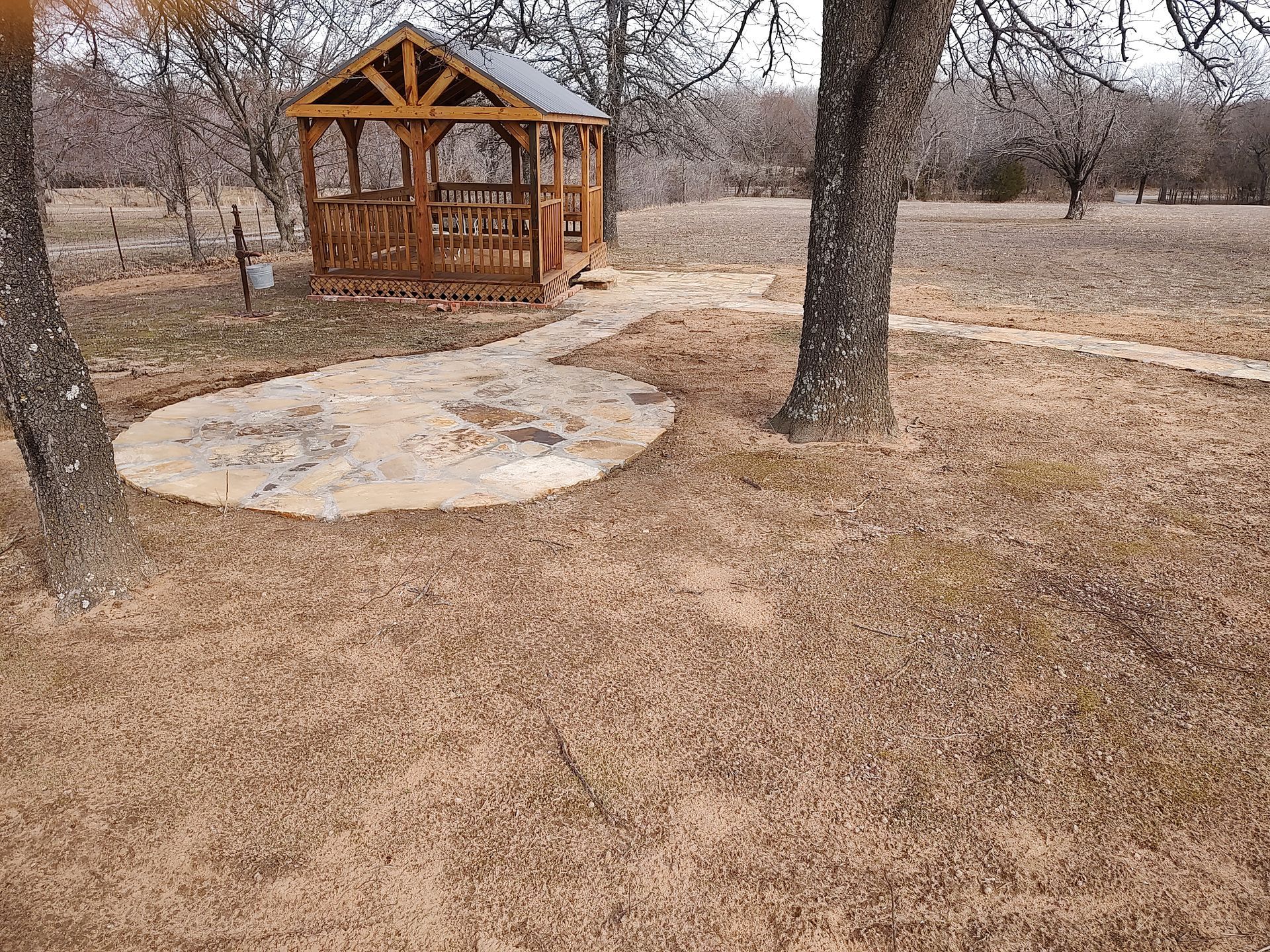 Wooden gazebo and stone fire pit in a grassy yard, trees in the background.