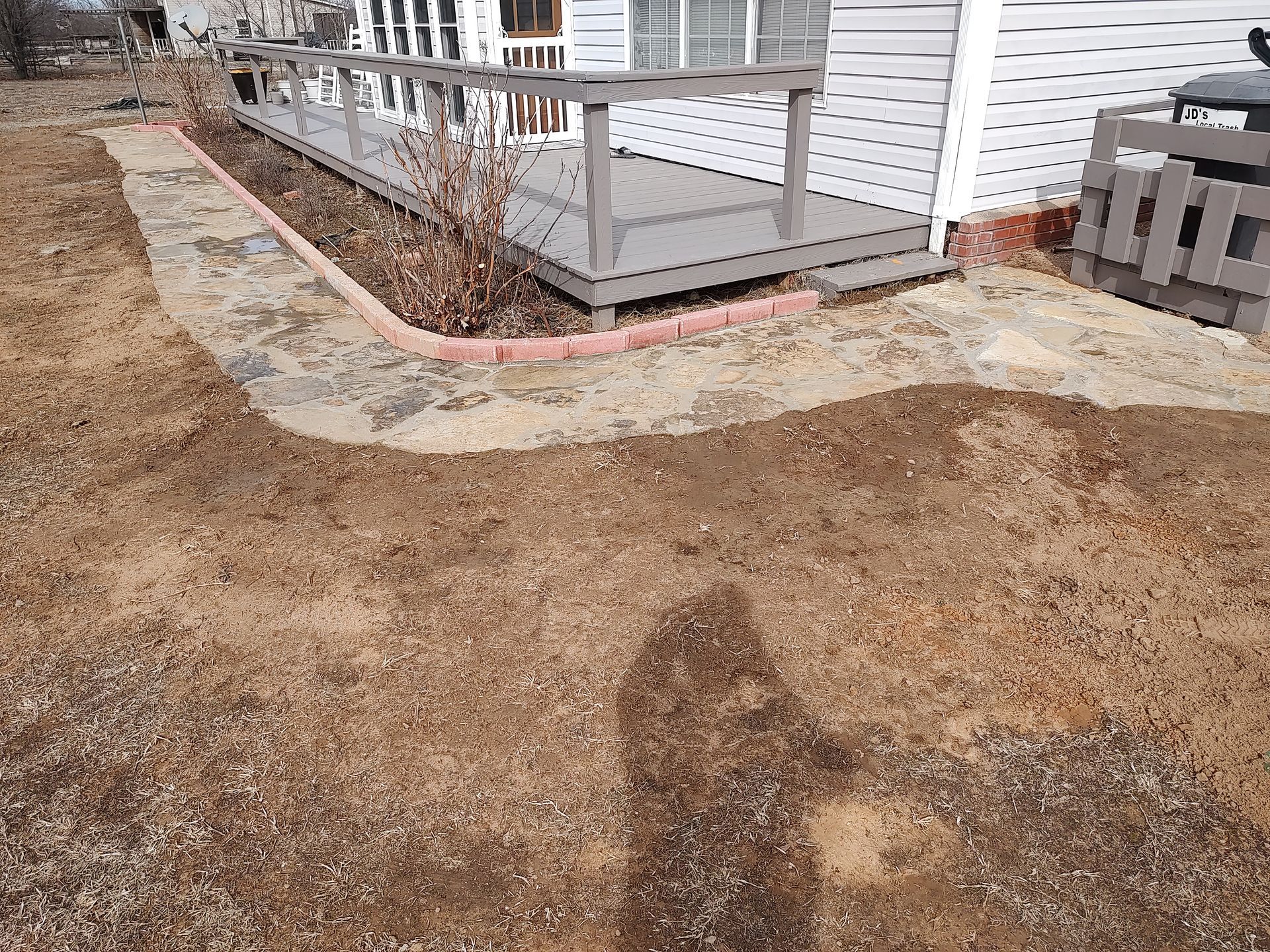 Brown yard with a brick-lined border next to a gray deck and white house.