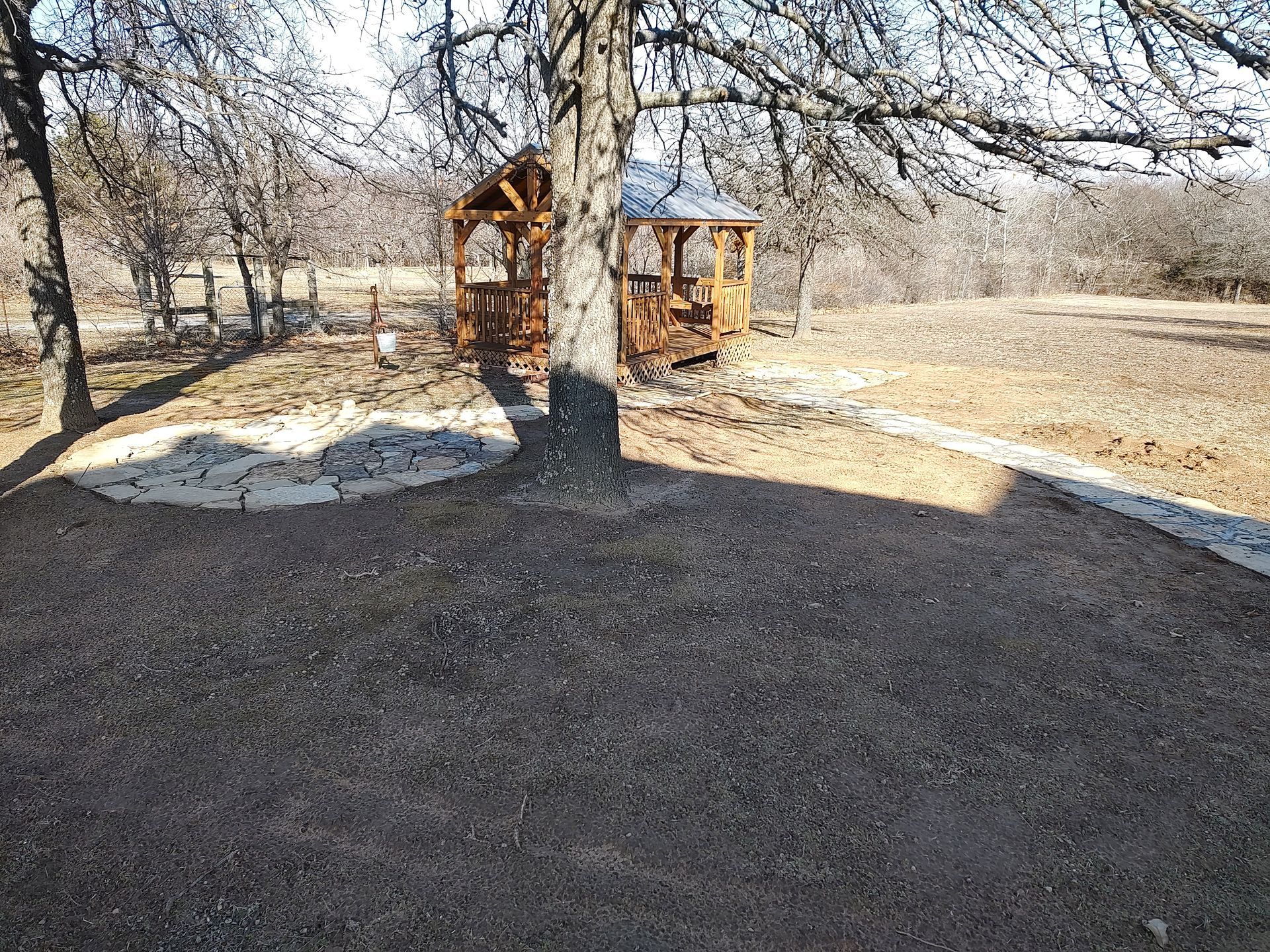Wooden gazebo in a brown, grassy area, under leafless trees. Sunny day.
