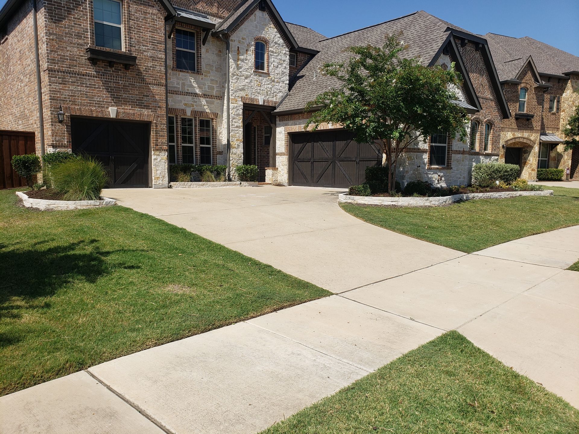 House with brick and light-colored stone facade; driveway and sidewalk in front. Green lawn and a tree.