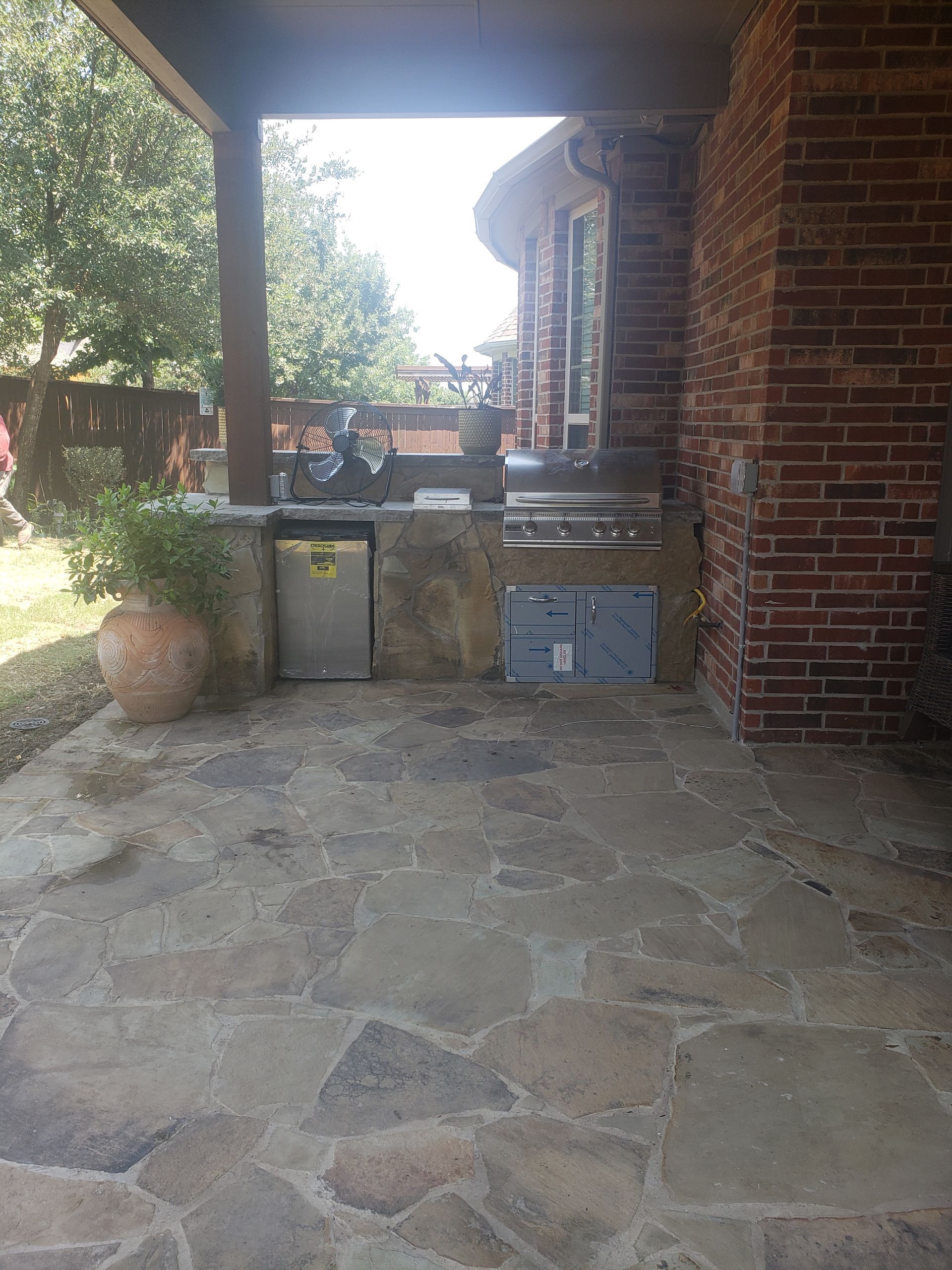 Outdoor kitchen on stone patio with grill, sink, and refrigerator. Red brick wall on right.