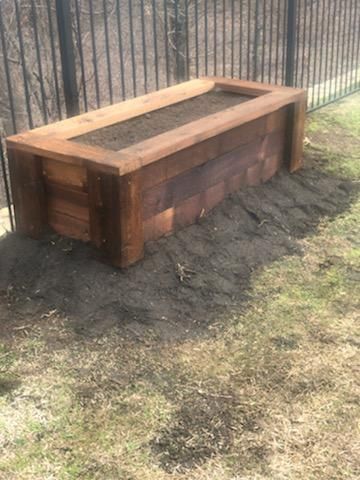 Wooden raised garden bed filled with soil, in a grassy outdoor setting, next to a fence.