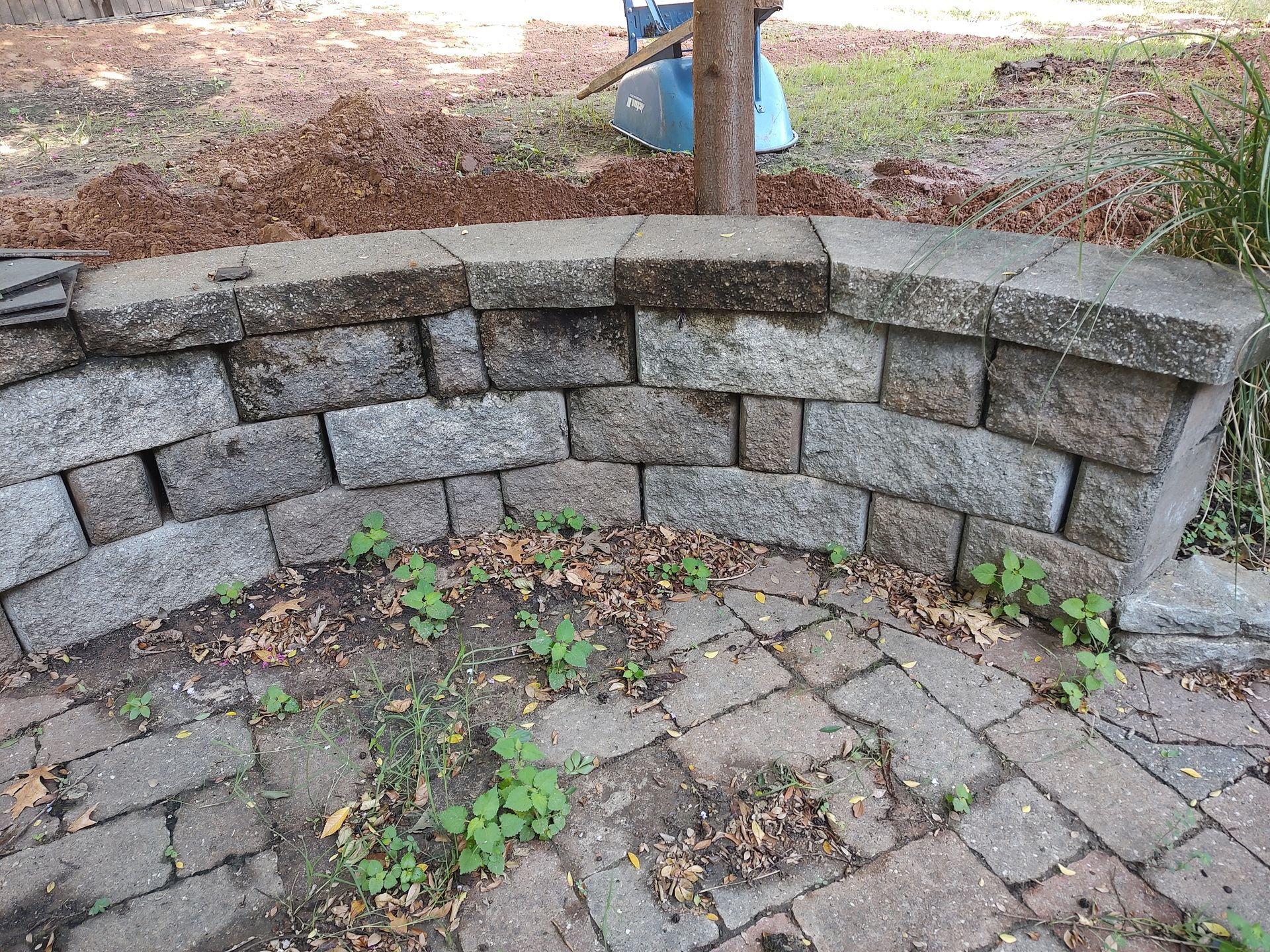 Stone retaining wall curving around a brick patio, with small green plants growing at its base.