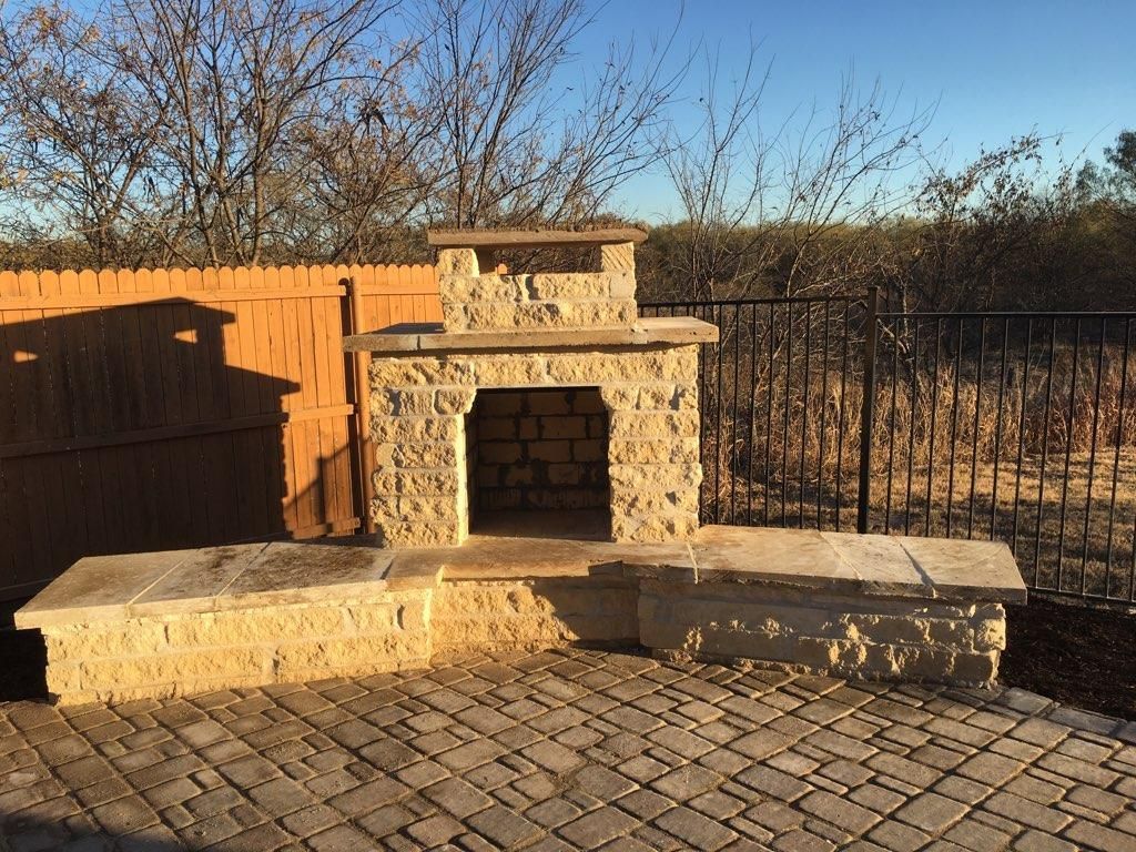 Stone outdoor fireplace and seating on a brick patio, against a wooden fence and a black metal fence.