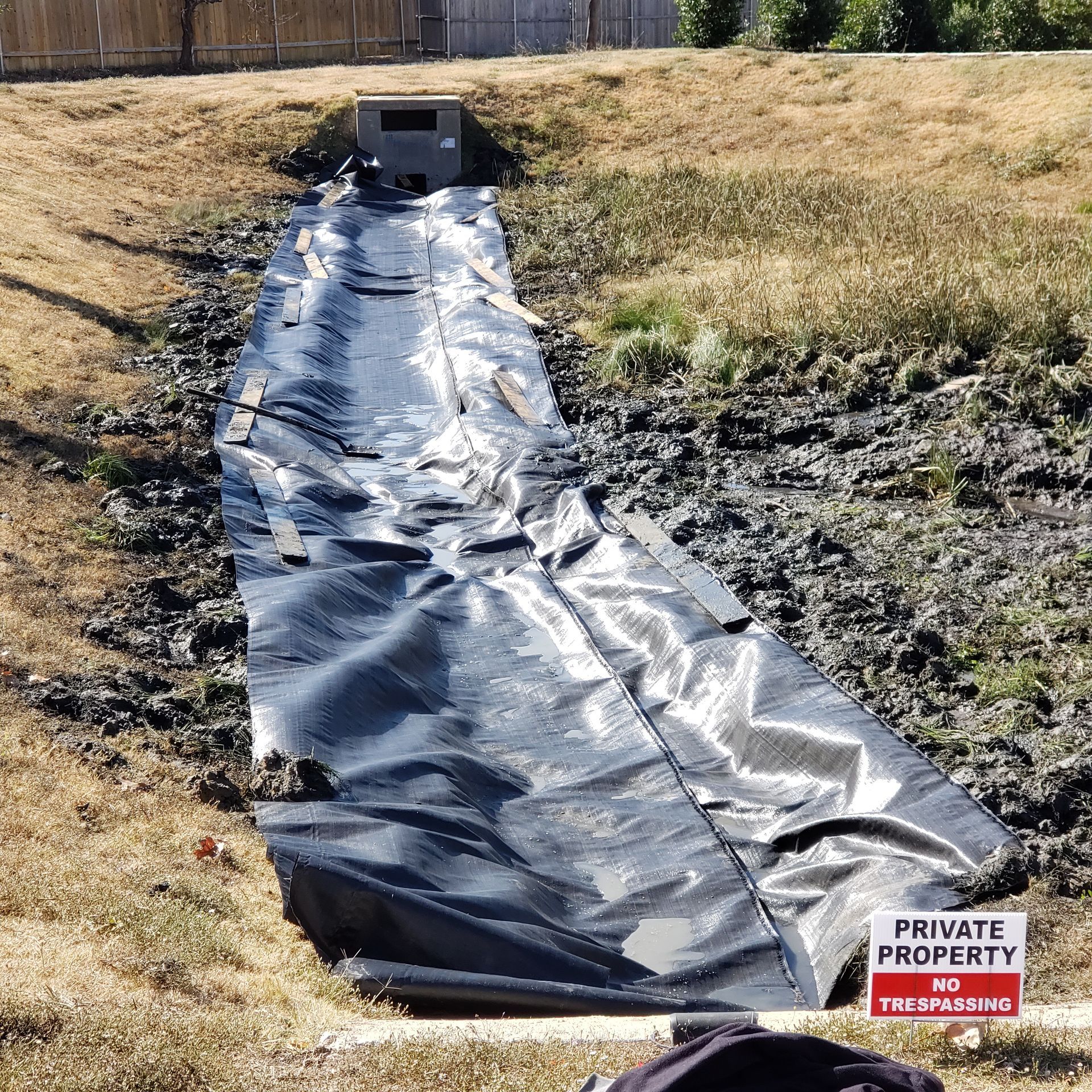 Black tarp covers a muddy drainage ditch leading to a concrete culvert. Brown grass and 