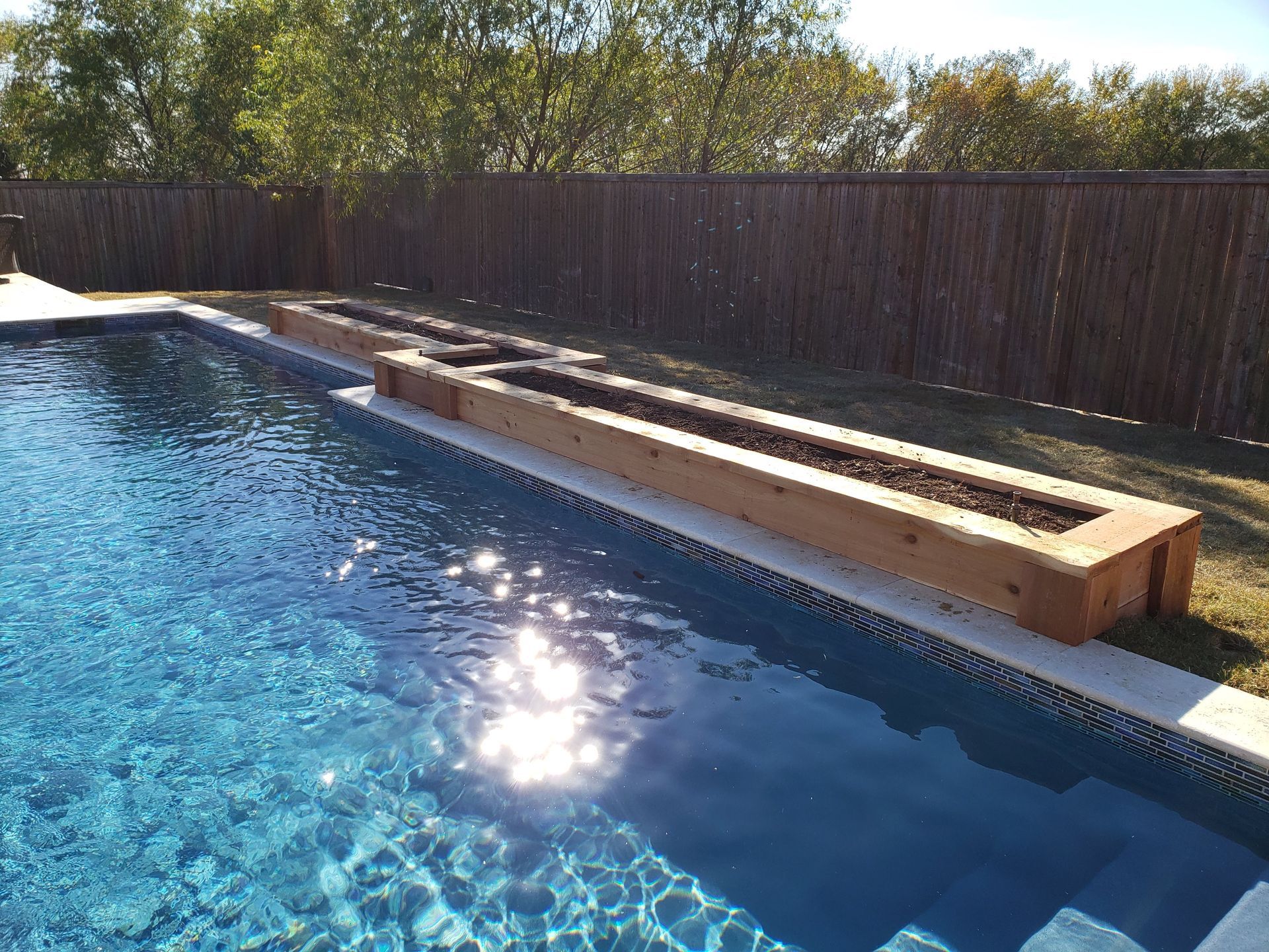 Swimming pool with wooden planters along the edge; a brown fence and grass in the background.