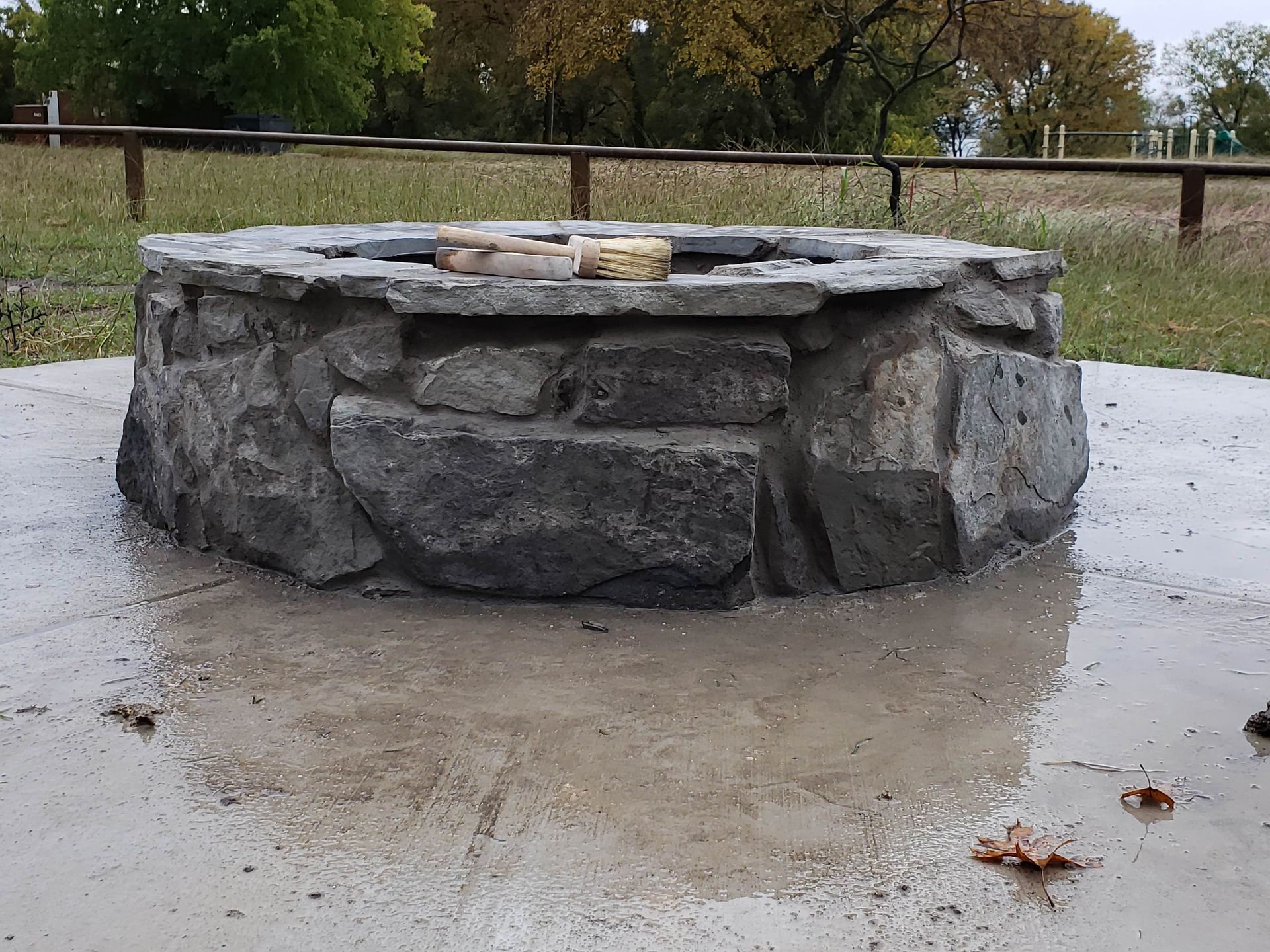 Stone fire pit on a wet concrete surface outdoors, with a fence and trees in the background.